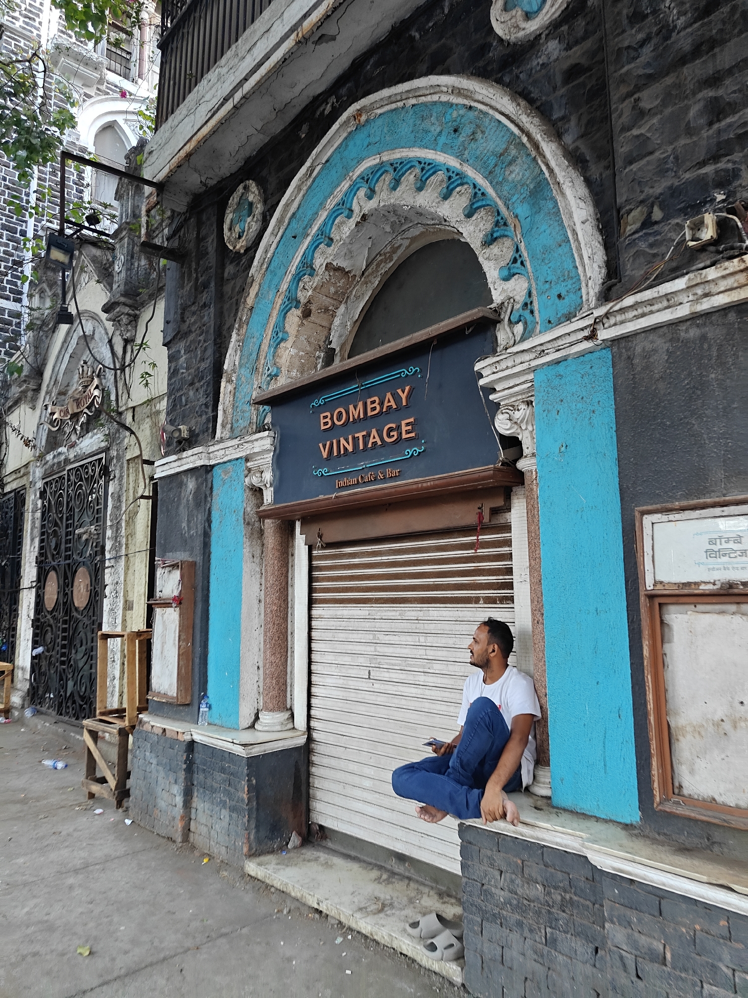 A man sits on a ledge outside a building in Mumbai, India. The building, "Bombay Vintage Indian Cafe & Bar", has an aged facade with cracked plaster and peeling paint in shades of blue, white, and brown. The man is barefoot, wearing jeans and a white t-shirt, and looks off to the left. A partially open roller shutter door serves as a backdrop. Nearby buildings are visible on the left, with a wooden structure and discarded bottles on the ground. A window to the right displays text in what appears to be Devanagari script.