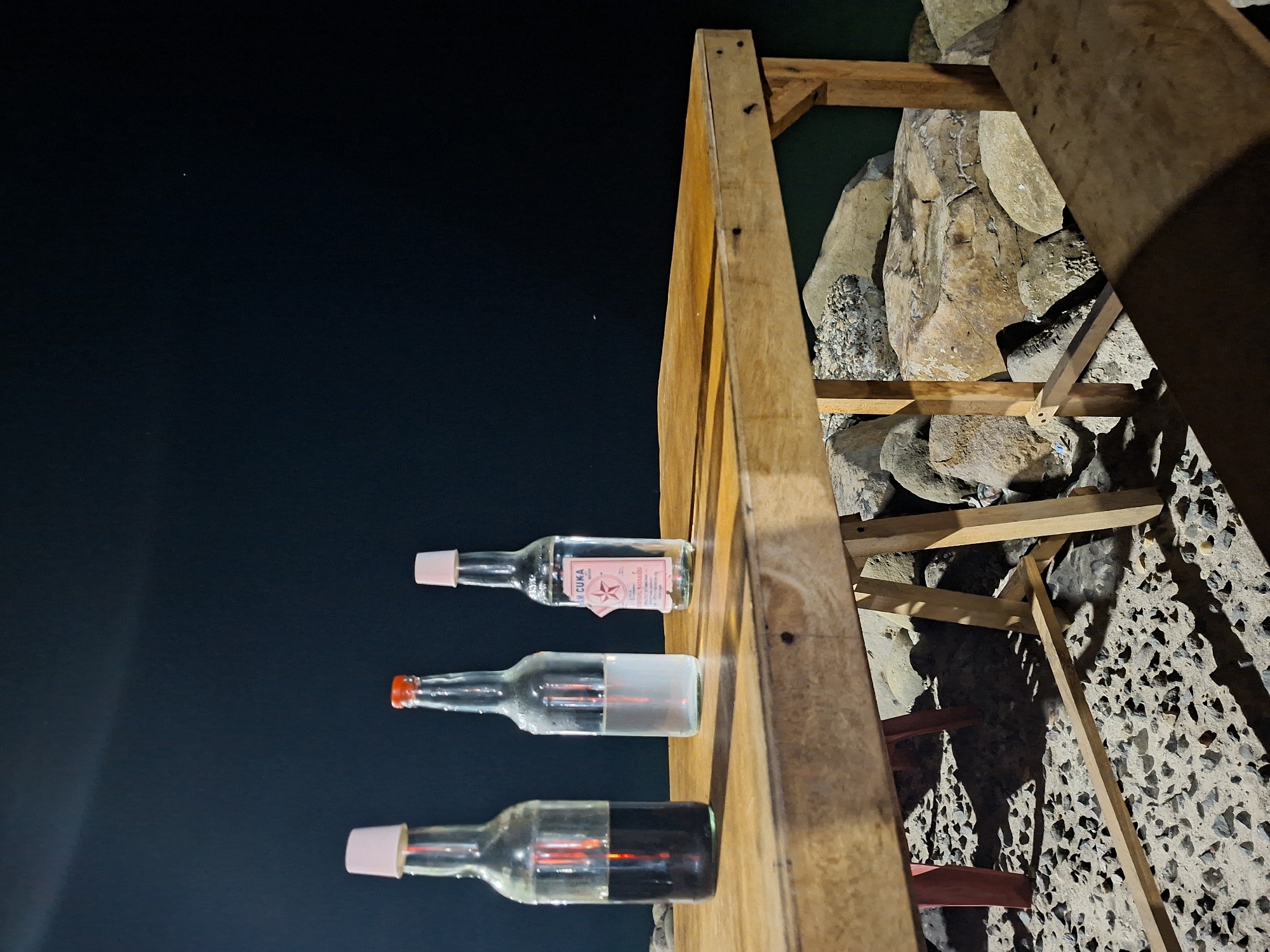 The image shows three bottles of liquids lined up on a wooden shelf against a dark, potentially nighttime, background. The top bottle is clear with a pink cap and has a label reading "CUKA" (vinegar), indicating the liquid is likely vinegar. The middle bottle appears to contain a clear liquid, and the bottom bottle contains a dark liquid, potentially soy sauce or another dark condiment. The shelf is made of unfinished wood and appears to be part of a larger structure with wooden supports and elements that are visible in the background. Behind the structure, there is a wall or slope made of rocks and stones. The setting seems to be outdoors, and the presence of vinegar bottles and the possible condiments suggests the location might be near a food stall or small local restaurant, possibly in the vicinity of Lhokseumawe, Indonesia. There are no people directly visible in the image.