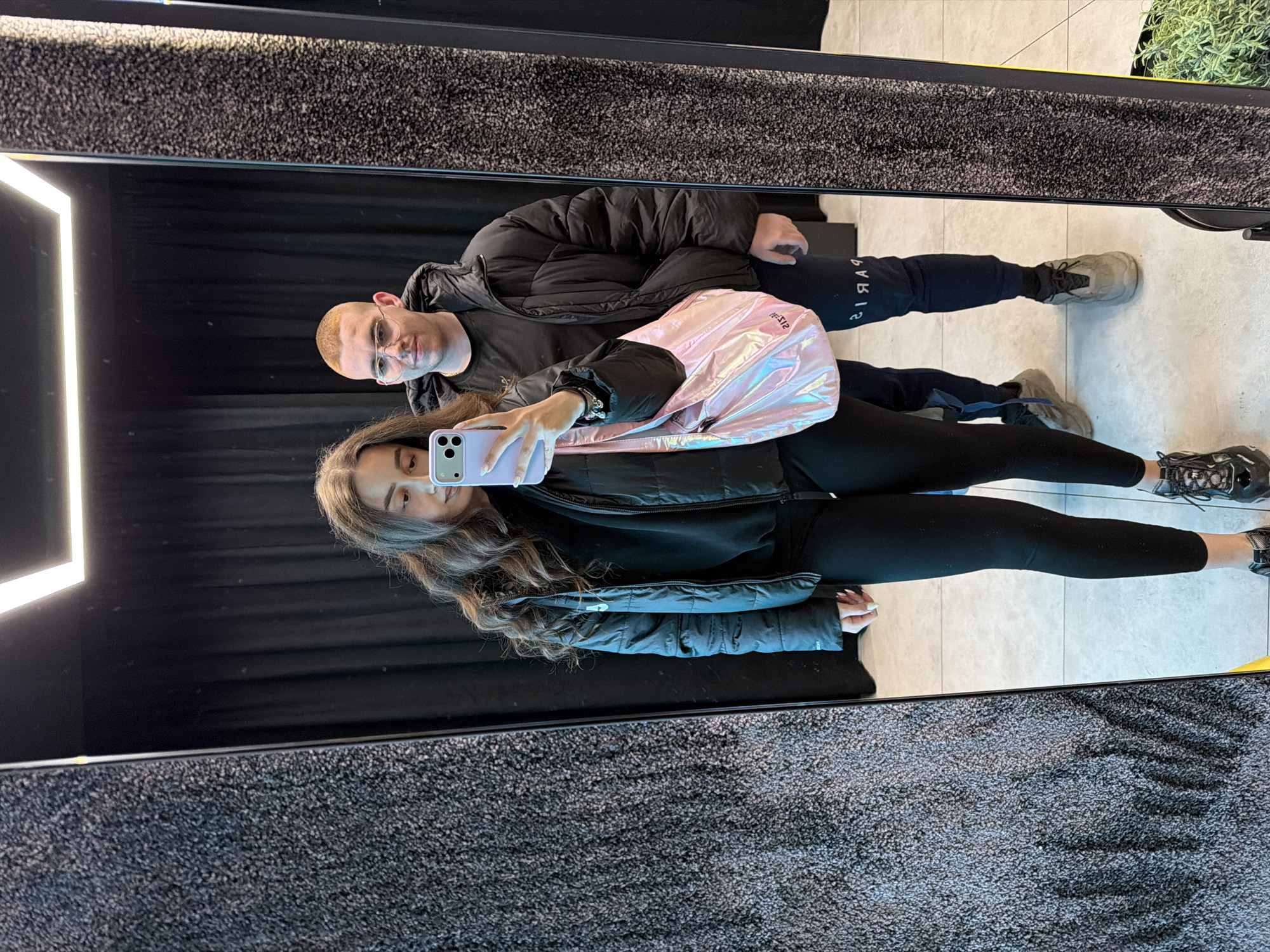 A young woman and man are captured in a mirror selfie. The woman, facing the camera, holds a light purple iPhone, and wears black leggings and a black puffer jacket. A shiny pink bag hangs on her shoulder. The man, with a shaved head and glasses, stands behind her, wearing a black puffer jacket and dark blue track pants with "PARIS" written on them. They stand on a beige tiled floor. Behind them is a dark curtain. The mirror frame appears to be textured. The location is a photo booth in Sofia, Bulgaria.