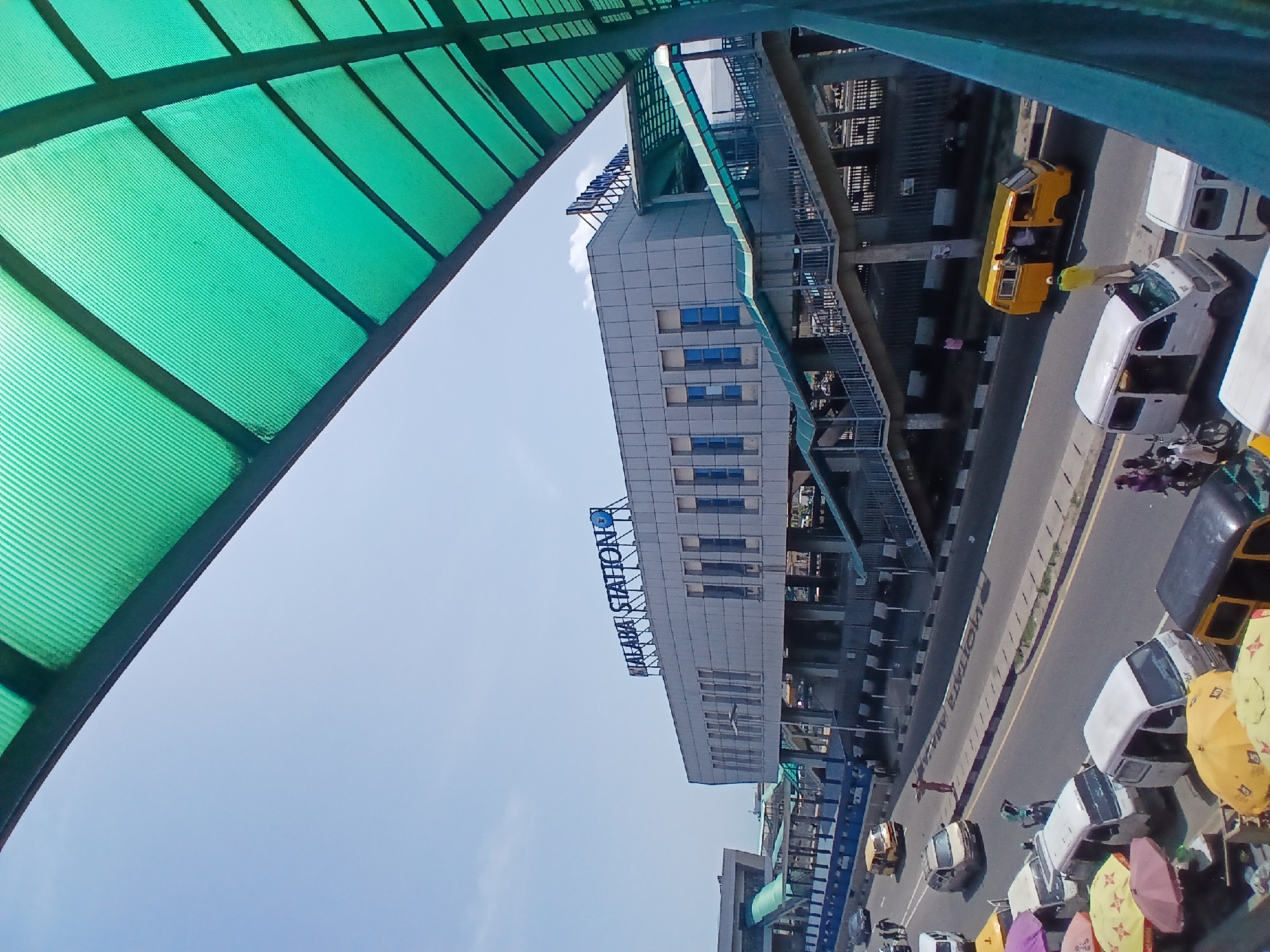 An outdoor, angled shot depicts the cityscape in Agani, Nigeria. The foreground includes a translucent green canopy. The Alaba Station building with the inscription "ALABA STATION" in blue letters is visible. Metal staircases are attached to the side of the station.
Vehicles like white and yellow vans are parked along the roadside. Colorful umbrellas are also visible near the vehicles. People are seen walking and engaging near the vehicles. The sky is clear and light blue.