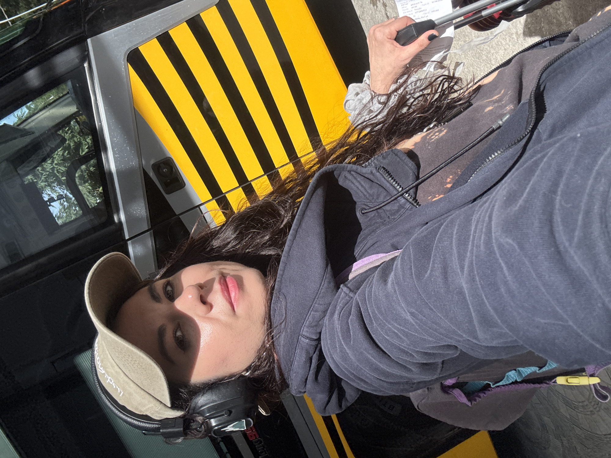 A woman wearing headphones and a baseball cap stands in front of a yellow and black striped bus. She is holding a suitcase and appears to be traveling. The lighting suggests it is daytime, and the environment appears to be an outdoor setting, possibly a bus station or a street in Da Lat, Vietnam, given the presence of trees and a building in the background, which are typical elements of Da Lat's landscape.
