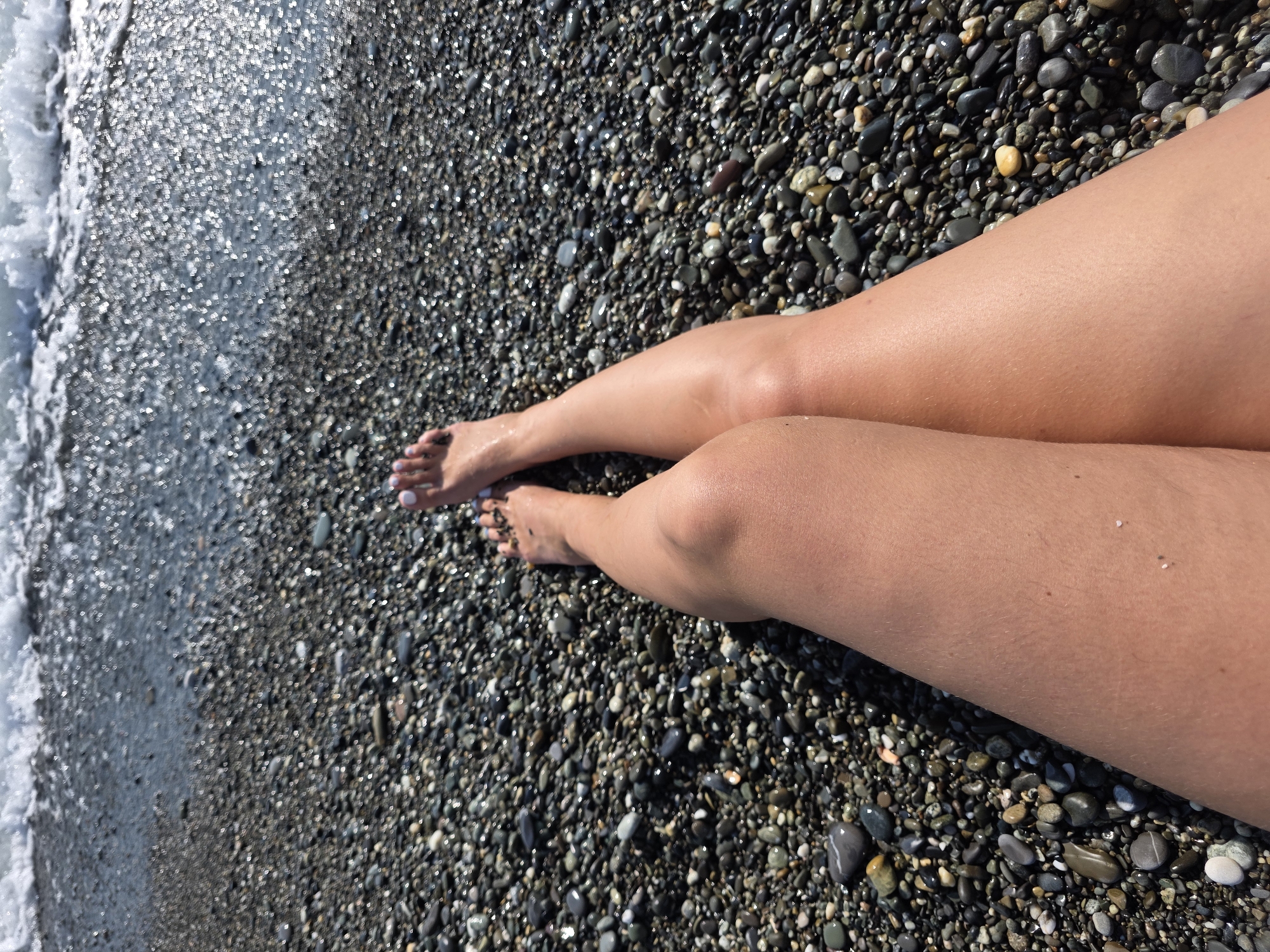 The image depicts a close-up, overhead view of a person's legs and feet on a pebble beach. The person is wearing white nail polish on their toes. The legs are tanned, suggesting a sunny day. The beach is covered in smooth, dark pebbles, many of which are wet and reflect the sunlight, creating a sparkling effect. To the left of the frame, a wave gently washes onto the shore, its white foam contrasting with the dark pebbles. The water appears clear. The scene is set at a beach, likely in Sochi, Russia, given the context. The overall impression is one of relaxation and enjoying a day by the sea. The time of day appears to be daytime, indicated by the bright sunlight and reflections. There is no visible text in the image.