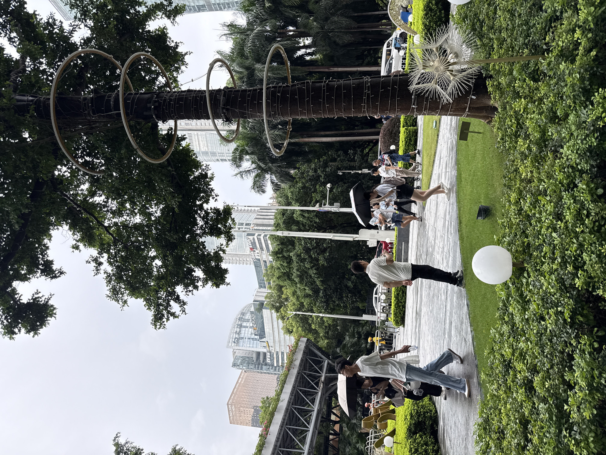 In Tianhe District, China, on an overcast day, a park scene unfolds with a group of pedestrians walking on a paved pathway bordered by manicured green bushes and grass. In the foreground, a spherical white light fixture is nestled amongst the foliage. To the right, a tall tree wrapped with string lights and adorned with three circular structures stands prominently. Beyond the trees, a modern cityscape rises with glass-fronted skyscrapers, including one labeled "GRAND PLAZA". Several palm trees are visible to the right of the central tree. People are dispersed across the scene; some walk with umbrellas, indicating recent or impending rain. One man in a white t-shirt and jeans walks towards the left, holding a clear bottle. Another man in a white polo shirt and dark pants walks to the right. A woman in black shorts and a white t-shirt, holding a black umbrella, walks with another individual. A woman with a light-colored umbrella is also present. A stroller is visible behind a woman in black. In the background, vehicles, including a white van, are parked. A low, angular metal structure with foliage on top is situated to the left, partially obscured by trees and people. A white pail rests on the grass near the pathway.