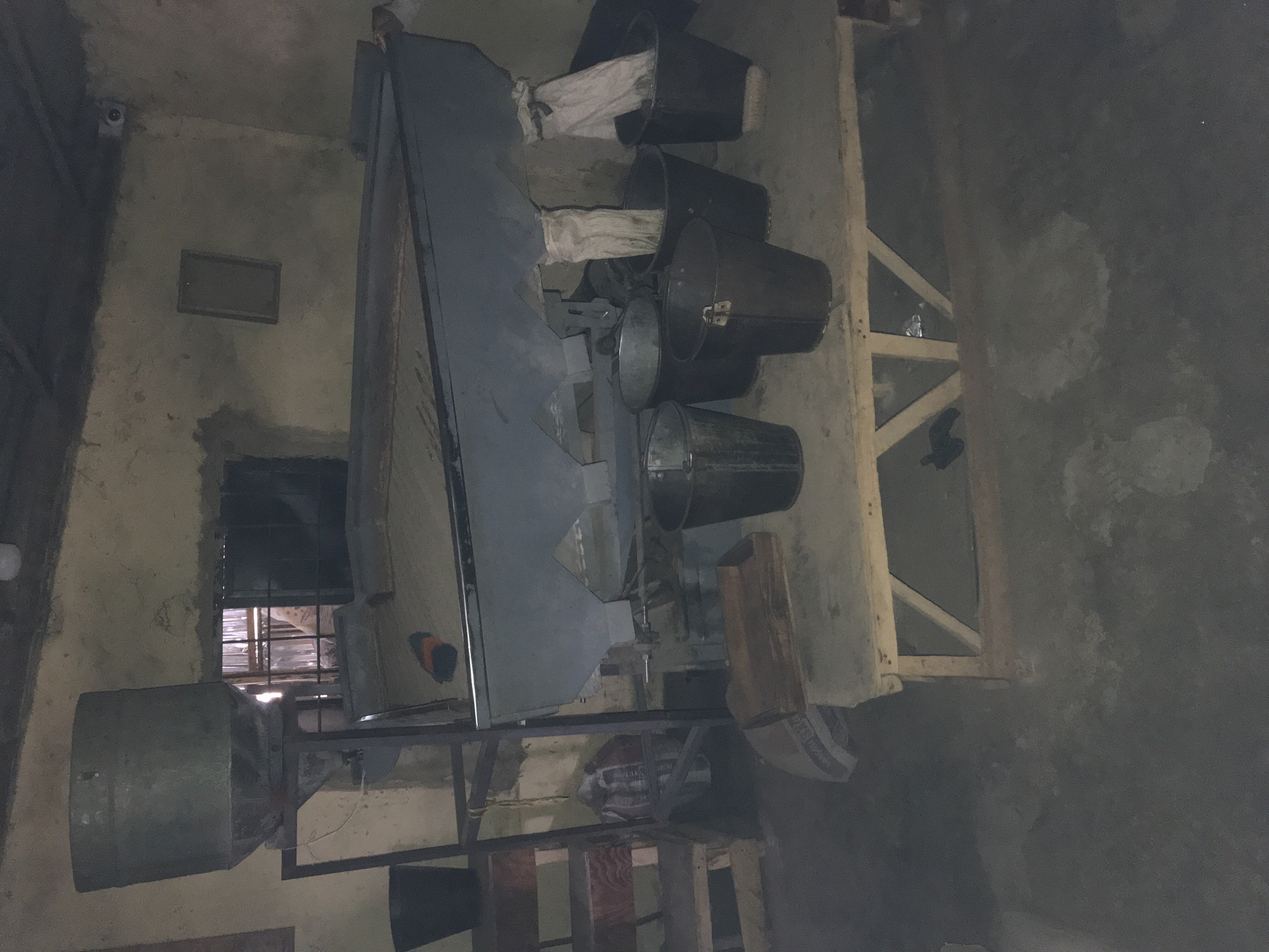 In Kabong, Nigeria, this image depicts a dimly lit, indoor industrial or workshop setting. A large, grey metal hopper or feeder is mounted on a metal frame at the upper left. Below the hopper, a dark grey, trough-like machine extends across the frame. Several black buckets are positioned beneath the trough, with some containing white fabric or material. To the left of the main machinery, a dark plastic bucket sits on the floor. A wooden structure with slatted shelves is visible behind the dark plastic bucket. A wooden platform, supported by angled legs, is situated in the foreground, holding some of the buckets. The walls appear to be rough and unfinished, with a window opening in the background. A small, dark object, possibly a piece of clothing or debris, is on the floor near the front of the trough. A security camera is mounted high on the wall to the right.