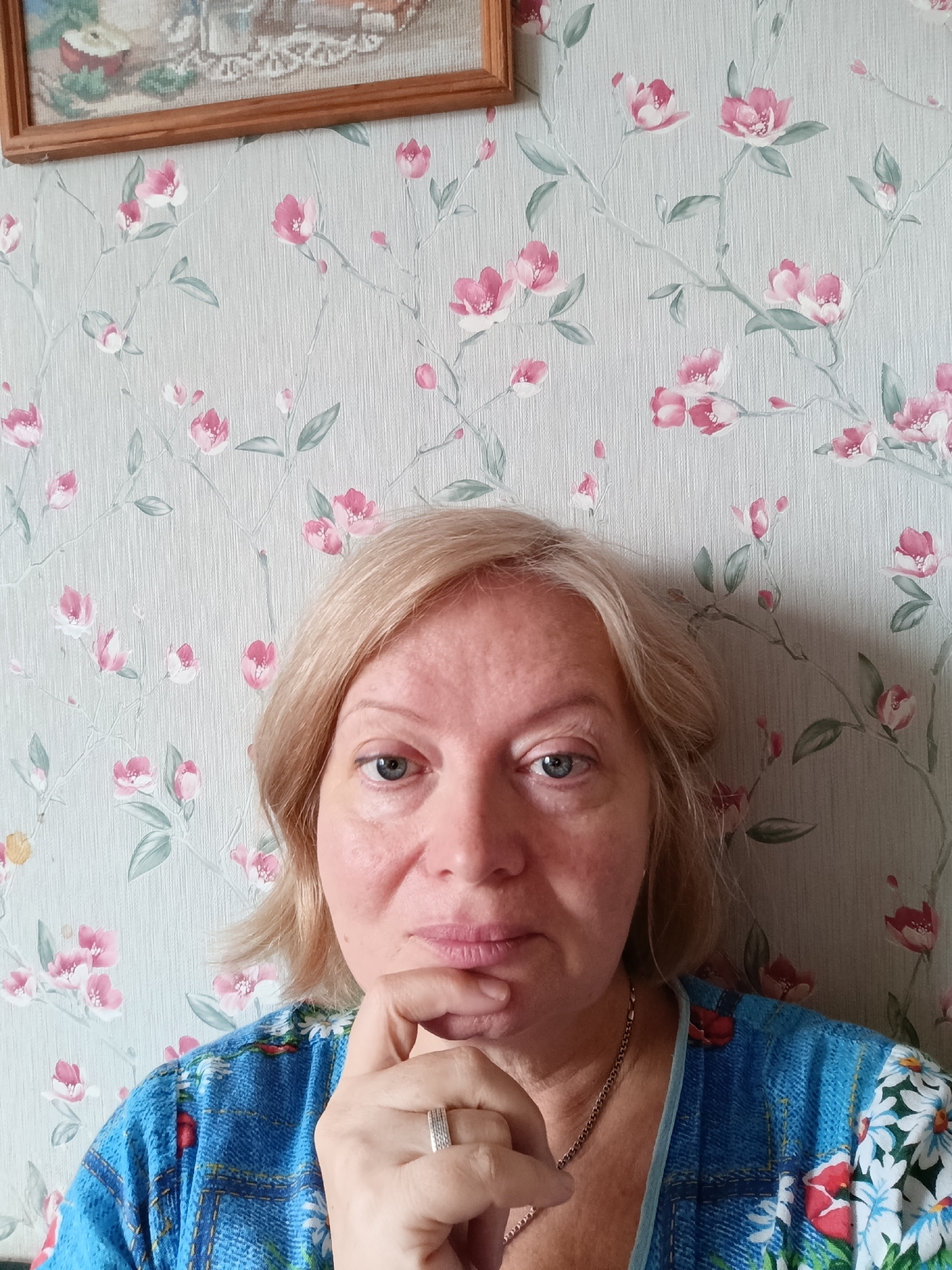 A medium, eye-level shot features a woman with fair skin and short, blonde hair. Her left hand is raised to her chin, suggesting a thoughtful pose. She is wearing a blue patterned top and a silver necklace. Her left ring finger has a silver ring.
The background consists of floral-patterned wallpaper in light blue with pink flowers and green leaves. A framed picture hangs on the wall near the upper left corner of the frame.
There is no identifiable information within the image to confirm the location in the city of Большой Букор, Russia.