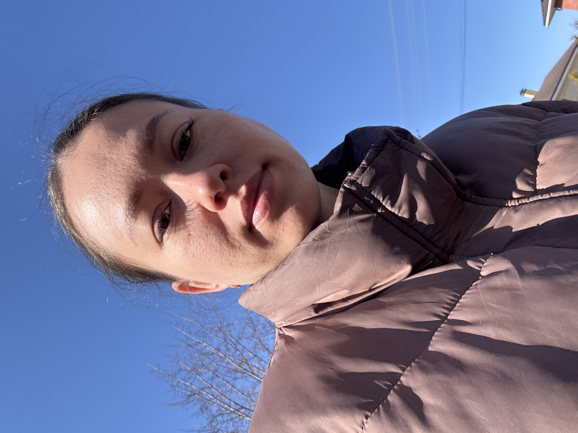 A woman is pictured from a low angle against a bright blue sky. She is wearing a brown puffer jacket with a high collar. Power lines are visible in the sky. Bare tree branches are in the lower left corner of the image. A roof and chimney are visible in the upper right corner. The woman appears to be outdoors. The location is likely Ферма, Russia.