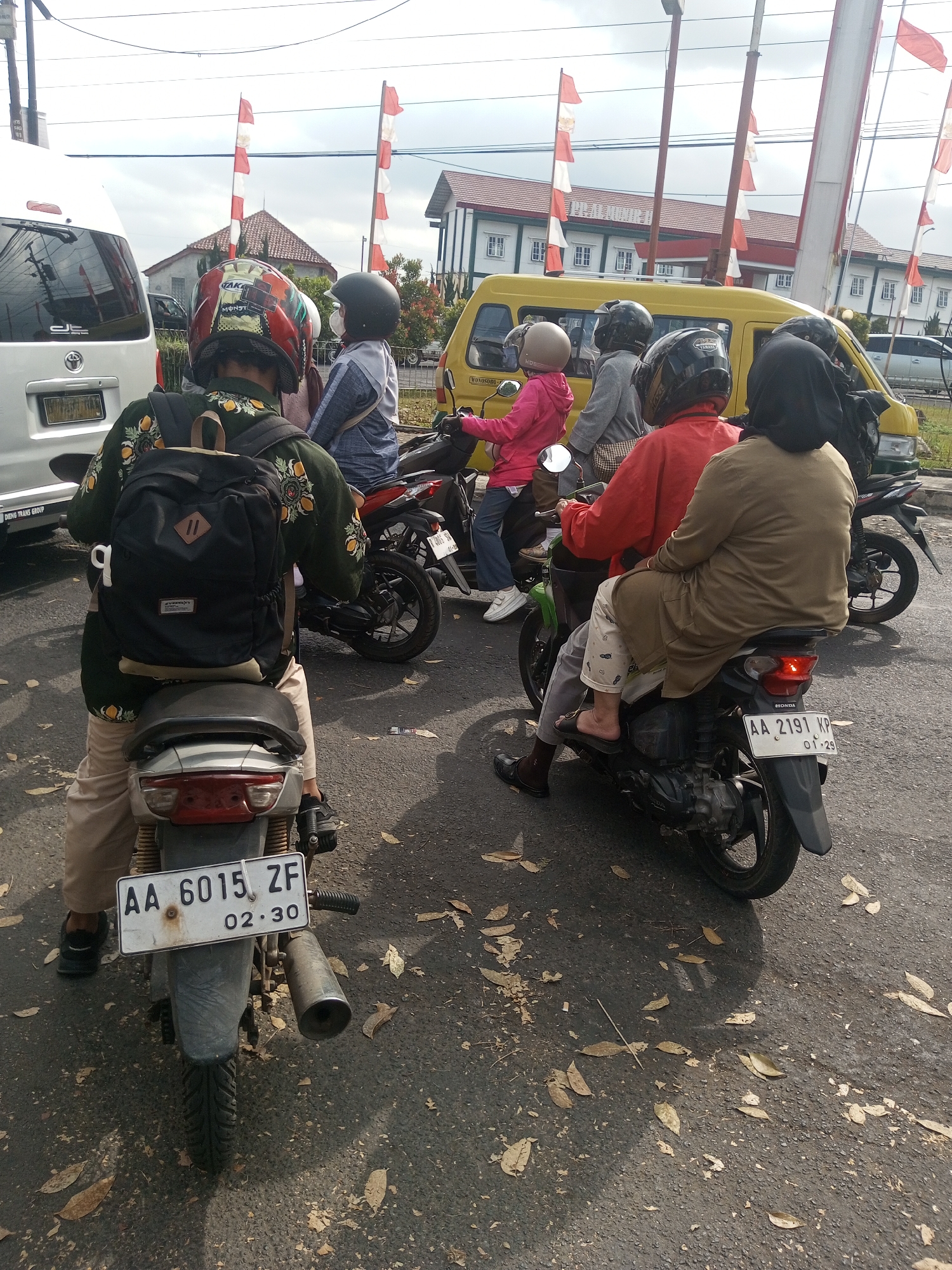 The image captures a street scene in Krasak, Indonesia, likely during the daytime given the illumination. Several motorcycles are visible, with multiple individuals riding on them. The foreground features a motorcycle from the rear, with a rider wearing a green patterned jacket and a black backpack. Another motorcycle is next to it, and further back, a yellow van is prominent, also with people on motorcycles in front of it. Red and white flags, possibly national flags, are displayed on poles in the background, suggesting a public area or event. The road surface is asphalt, with fallen leaves scattered across it. The environment appears to be an urban or semi-urban setting with buildings and utility poles visible in the distance. The atmosphere suggests a typical street scene in Indonesia, with a common mode of transportation being motorcycles.