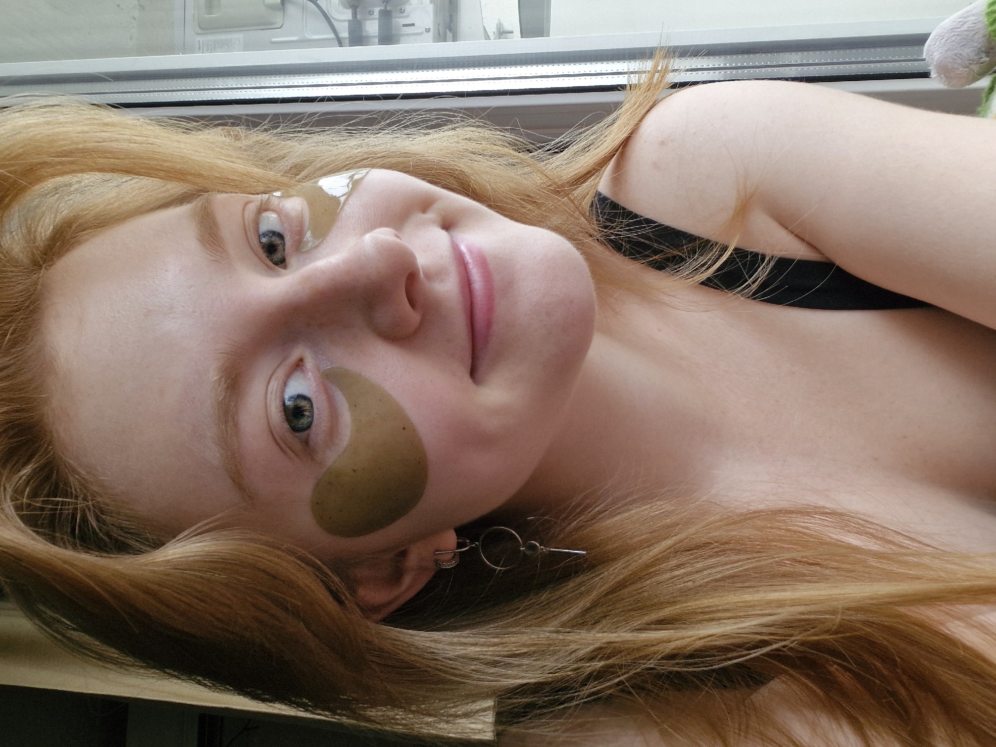 A fair-skinned woman with long, reddish-blonde hair is lying down, facing the camera. She has a cosmetic eye patch under each eye and a silver earring in her left ear. She is wearing a black tank top. She has blue eyes, freckles, and is smiling slightly with pink lipstick. The background is a window frame with what appears to be a cable and a small stuffed animal visible through the glass. The exact location in Yekaterinburg, Russia, cannot be determined from the image.