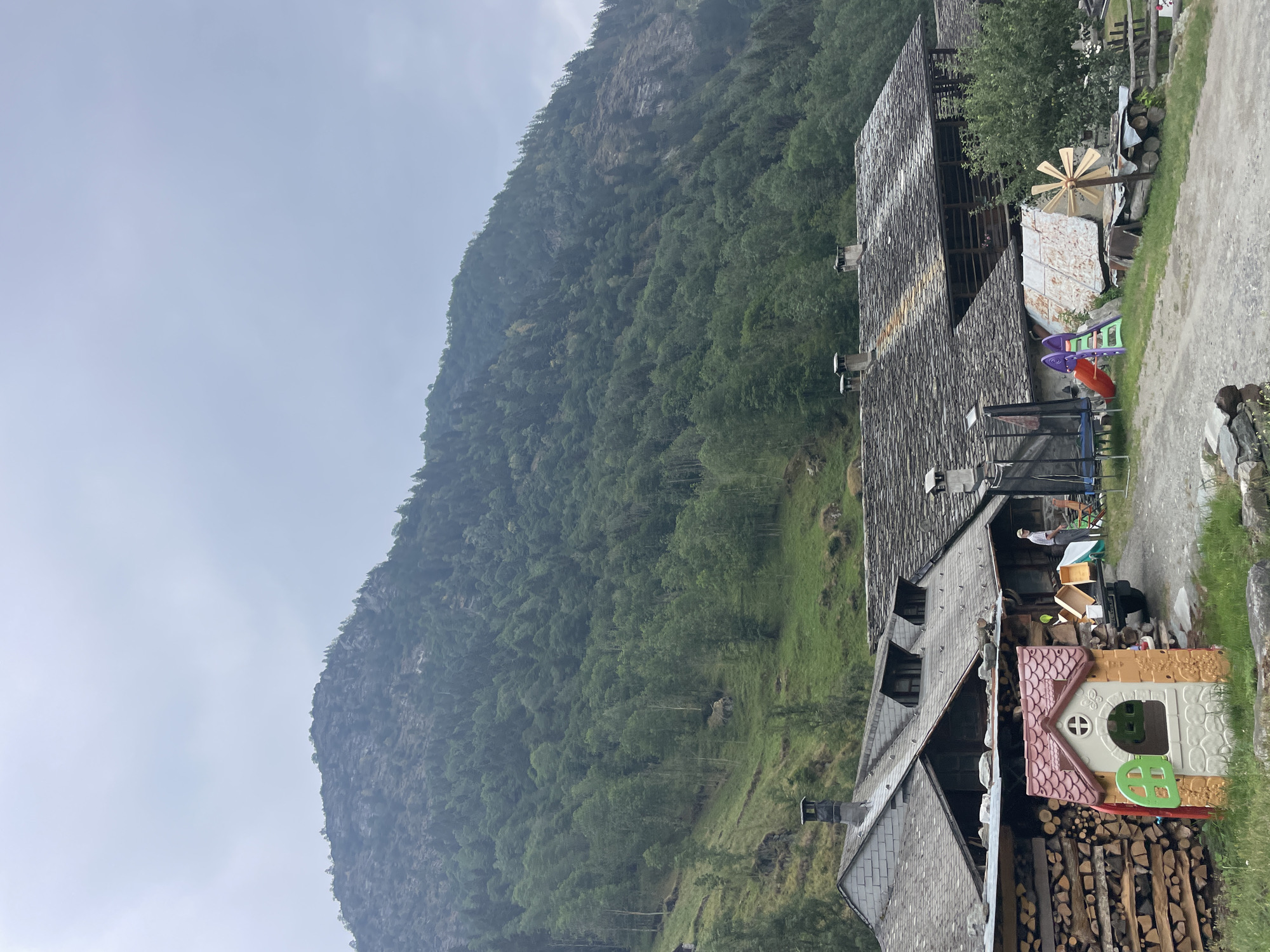 The image depicts a rural scene in Alagna Valsesia, Italy, dominated by a steep, tree-covered mountain under a hazy sky. In the foreground, several rustic stone-roofed buildings are clustered together. To the left, a large pile of neatly stacked firewood is visible. Adjacent to it is a small, brightly colored plastic playhouse. Further to the right, a trampoline and a slide are set up, indicating a play area. A person wearing a light-colored shirt and hat stands near a pickup truck with its bed open, appearing to be engaged in some activity. The ground is a mixture of gravel and grass. The architecture of the buildings, with their stone roofs and wooden elements, suggests a traditional alpine style. The overall atmosphere is one of a quiet, working mountain village or farmstead.