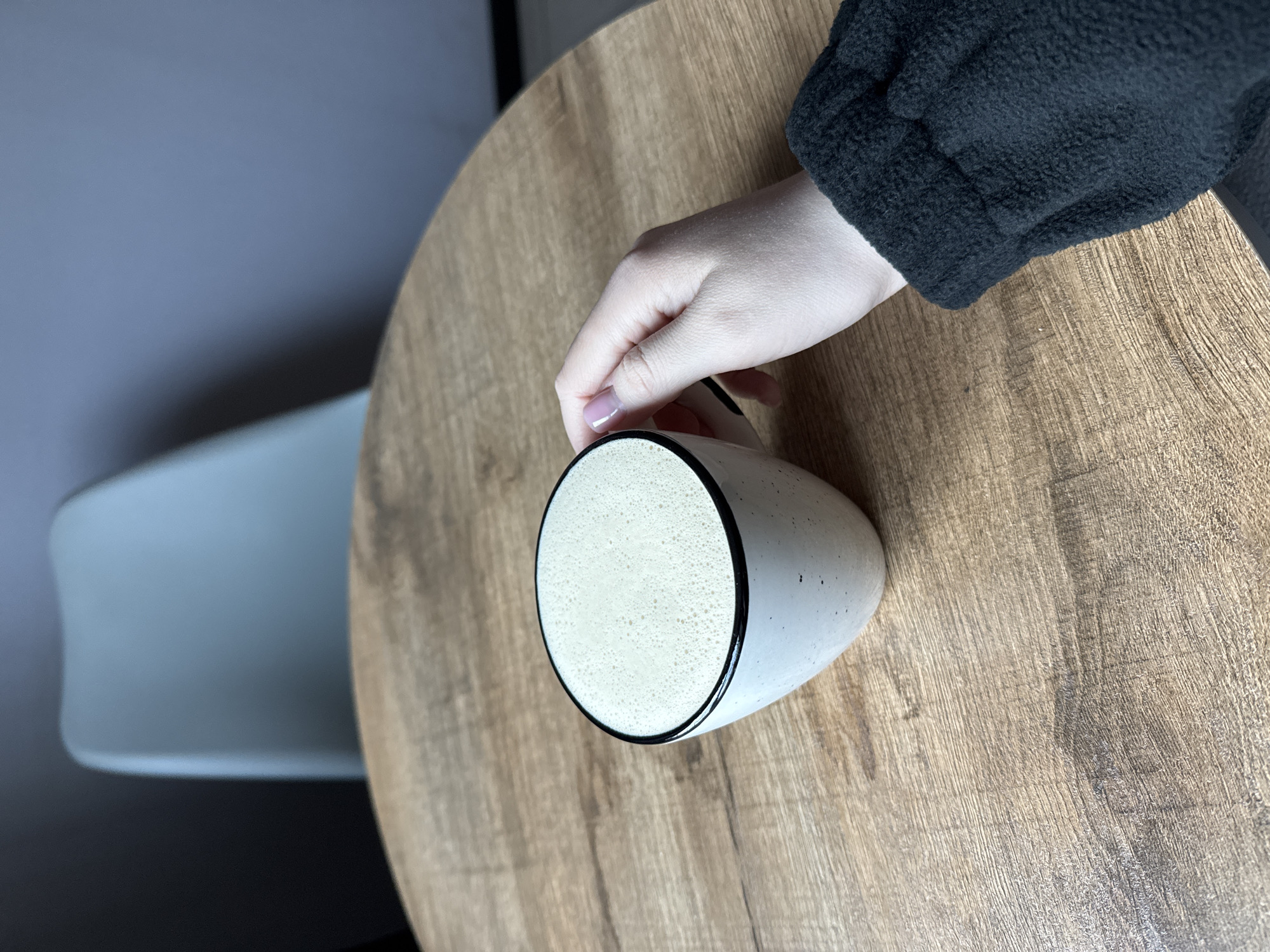 A high-angle shot depicts a white mug with a black rim, filled with a light-colored, foamy beverage, resting on a round wooden table. A person's hand with a light skin tone and light pink nail polish is placed on the mug. The person wears a black, textured sleeve. A light gray wall is visible in the background, along with the blurred image of a white chair. The location is Unknown, Unknown.