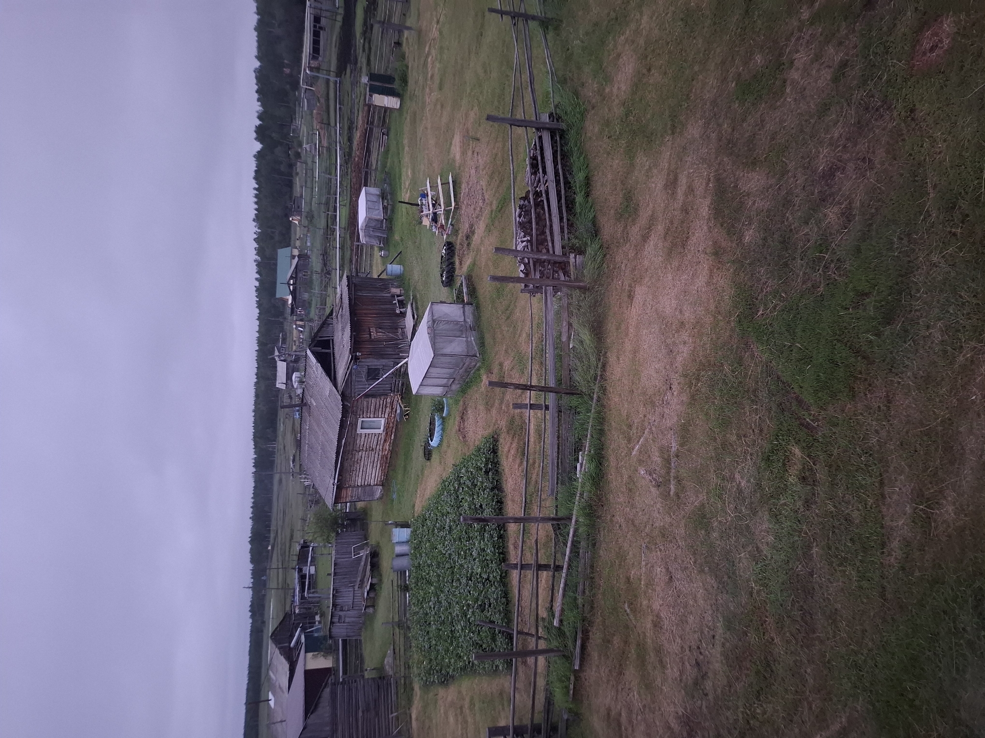 The image depicts a rural village scene in the city of Kyuelyany, Russia, under an overcast sky. The foreground features a wooden fence and dry, patchy grass. Beyond the fence, a large, well-tended garden bed is visible, bordered by more dry grass. To the right of the garden are several structures, including a wooden house with a window, a greenhouse, and what appears to be a woodpile. In the background, more wooden houses, sheds, and agricultural outbuildings are scattered across the landscape, with a line of trees forming the horizon. The overall impression is one of a remote, self-sufficient settlement.