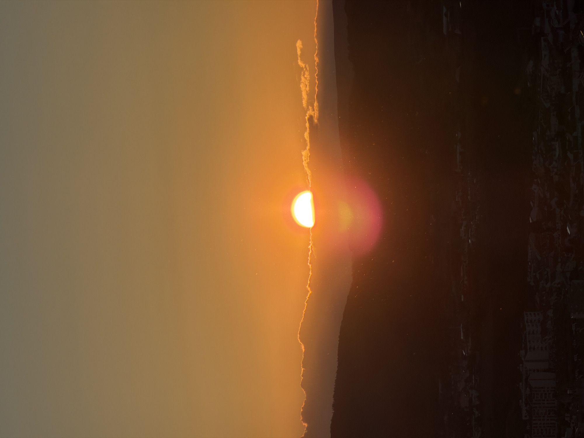 The image shows a sunset viewed from Bragança, Portugal. The sun is half-obscured by the edge of a city silhouette, casting a golden light in the sky to the left. The silhouette, with numerous rectangular structures, occupies the right portion of the image. The sky gradient shifts from a dark orange near the city skyline to a lighter yellowish hue in the left third of the image. There are small clouds on the skyline. The photograph is taken vertically.