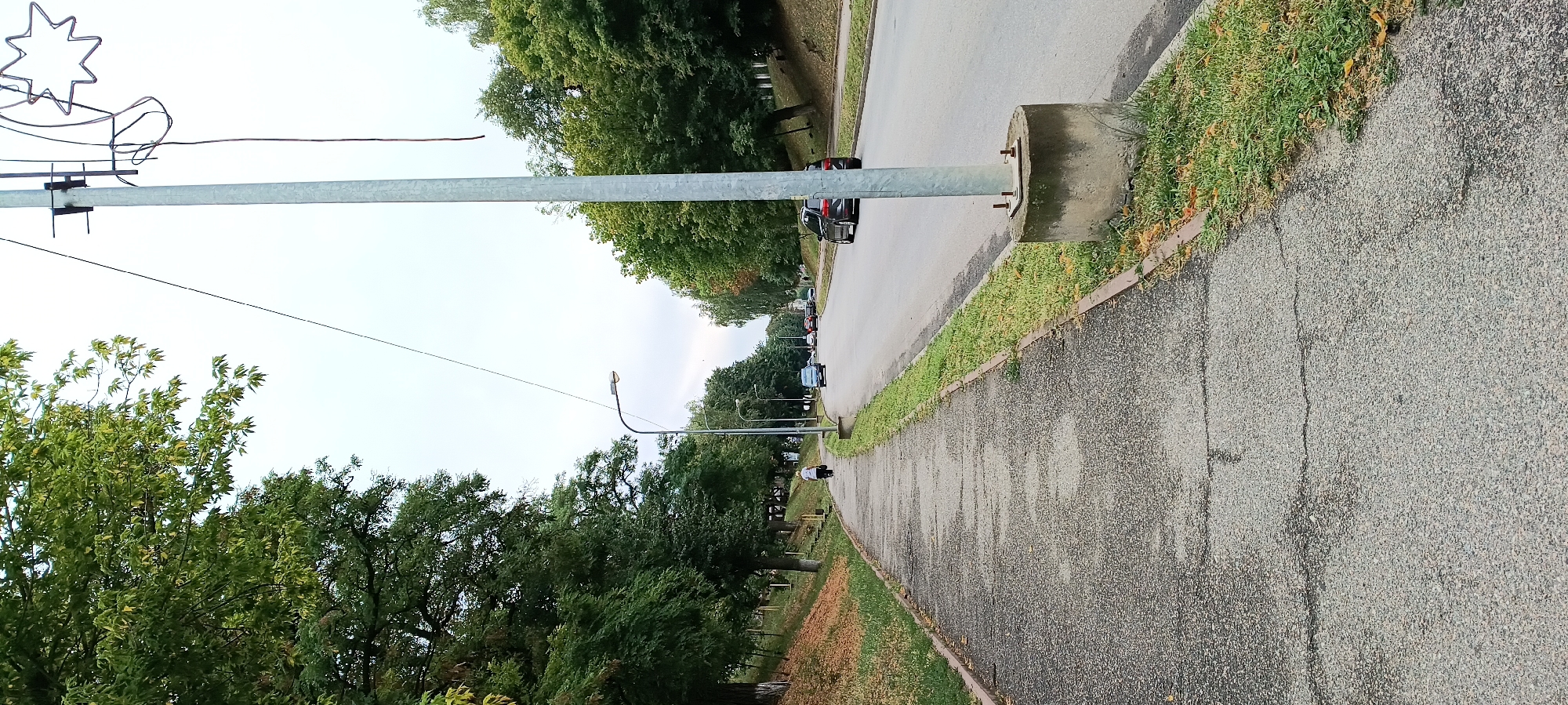 The image shows a street scene in Kamianka, Ukraine. A paved sidewalk runs along the left side of the image, bordered by a grassy area and trees. A lone person is walking away from the camera on the sidewalk. The street itself is asphalt and has two lanes of traffic. Several cars are visible driving on the street, and trees line the right side of the road. A utility pole with a street lamp and some decorative star-shaped lights is located on the right side of the sidewalk, near the edge of the road. The sky is overcast.