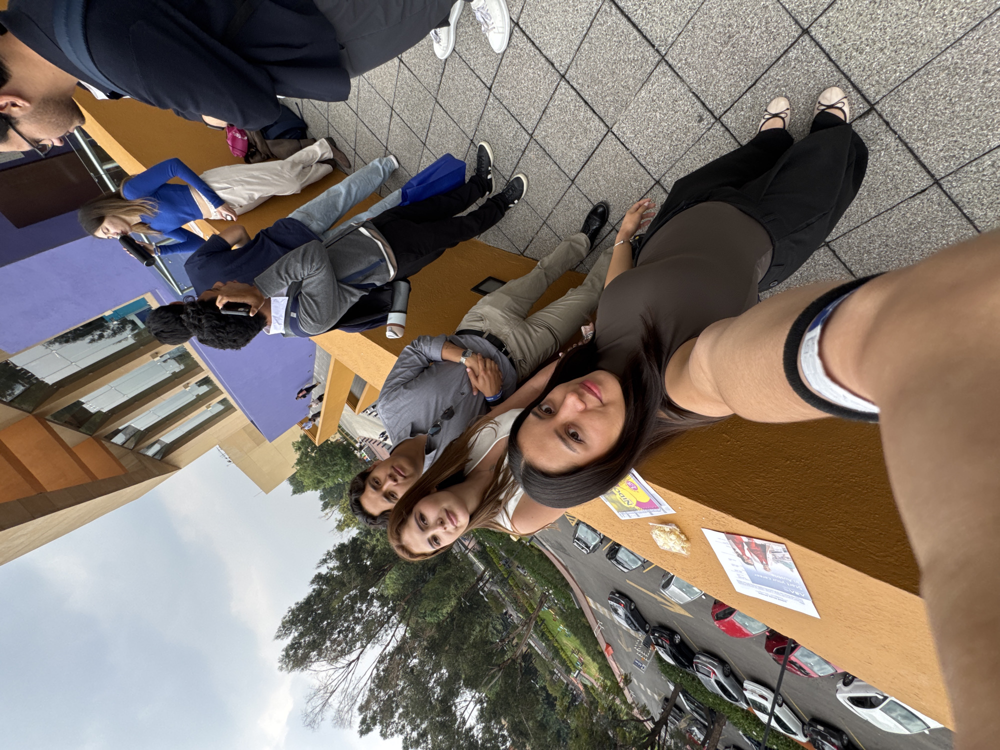 A selfie shows a group of people posing together in Mexico City, Mexico. The group is sitting on a ledge overlooking a street with parked cars and trees. There are at least eight people in the image, with three closest to the camera. The people are dressed in casual clothing, and they appear to be relaxed and smiling. There are flyers and some popcorn on the ledge. The architecture visible in the background is modern, with orange and purple buildings. The sky is cloudy.