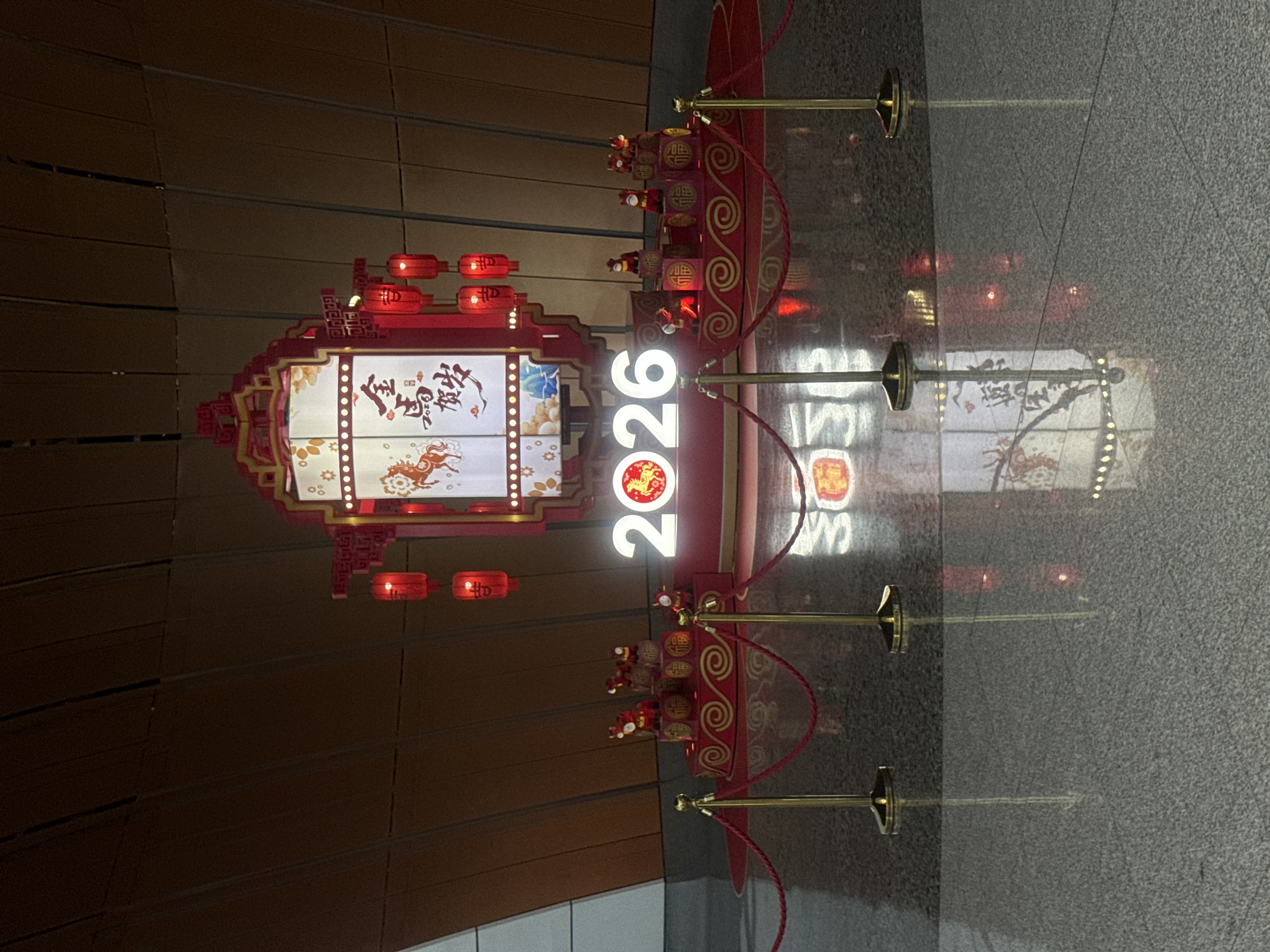 In Guangyang District, China, a festive Chinese New Year display features the prominent illuminated numbers "2026" in white, with the "0" in the center replaced by a red circle containing a golden horse. This display is set against a backdrop of a large, ornate lantern-like structure adorned with red lanterns and Chinese characters. Below this, a red platform showcases several small, red figurines, possibly representing zodiac animals, placed on tiered steps decorated with swirling patterns and Chinese characters that translate to "fortune." Gold stanchions with red ropes cordon off the display area. The entire scene is reflected on the polished, dark grey floor, creating a symmetrical visual. The background consists of curved, wood-paneled walls.
