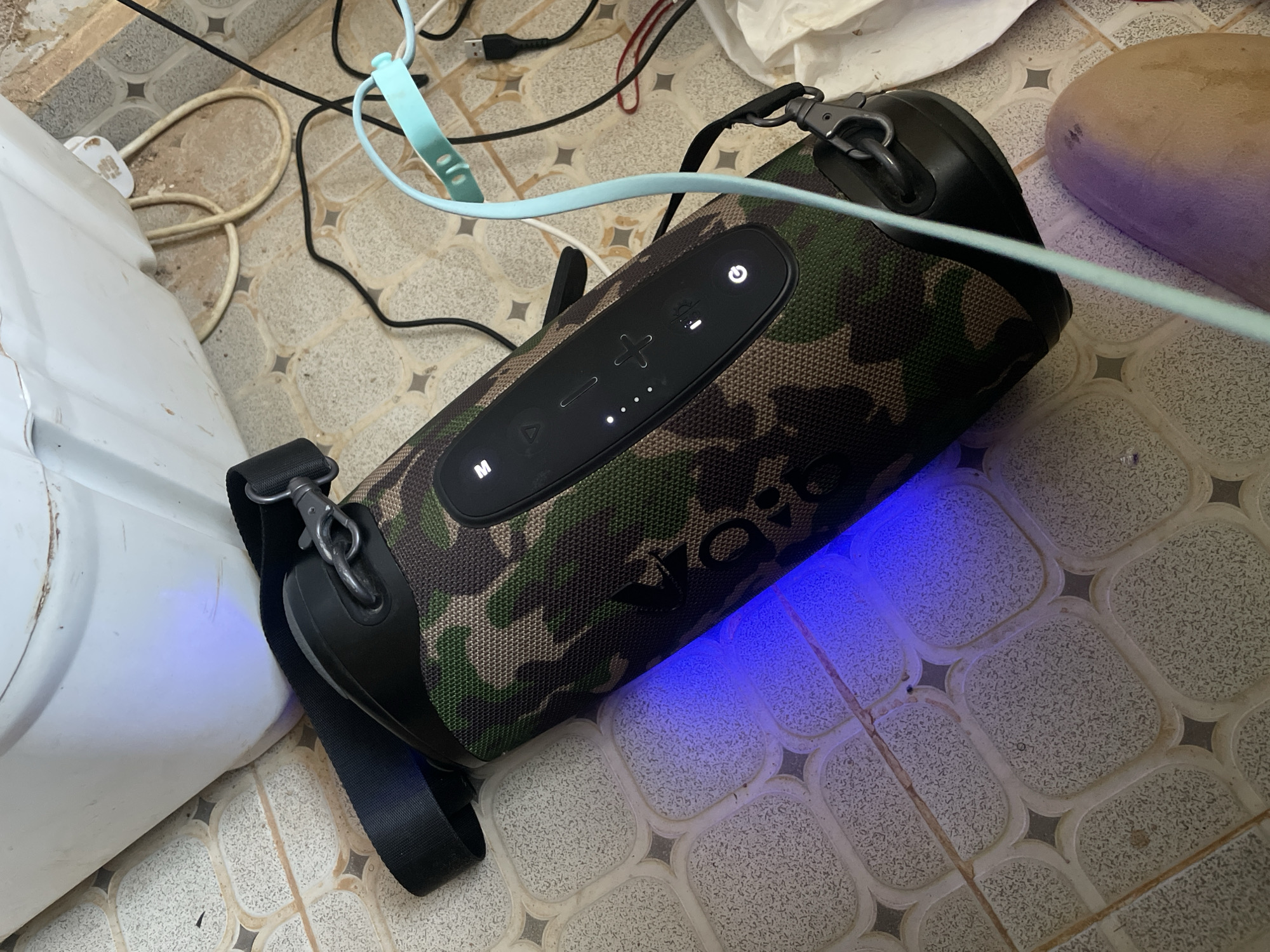 The image shows a camouflaged portable speaker on a patterned tile floor. A white plastic container is partially visible to the left of the speaker. Various cables and wires, including a light blue cable, are tangled around the speaker and container. A tan object, possibly fabric, is partially visible to the right. The speaker emits a blue light. The location is Rafin Bauna, Nigeria.