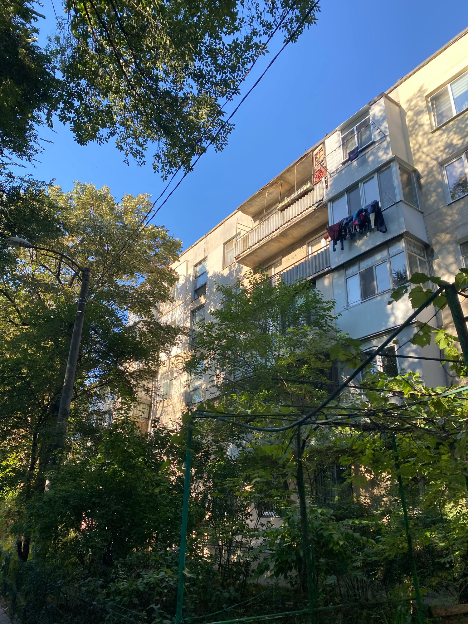 An upward-angle shot captures a building in Chișinău, Moldova, bathed in the warm light of a sunny day. The building, likely an apartment block, shows signs of everyday life with laundry hanging from a balcony on the third floor. The laundry includes a mix of dark and colored garments. A light-colored wall contrasts with the clear blue sky, with the shadows of trees dappling the building's facade. Lush green foliage from nearby trees partially obscures the view of the building, suggesting an urban environment with significant greenery. A street lamp stands to the left, and a metal trellis covered in vines is visible in the foreground, adding to the impression of a residential area. The overall scene depicts a common urban vista in Chișinău, showcasing the integration of residential architecture with natural elements and the daily activities of its inhabitants.
