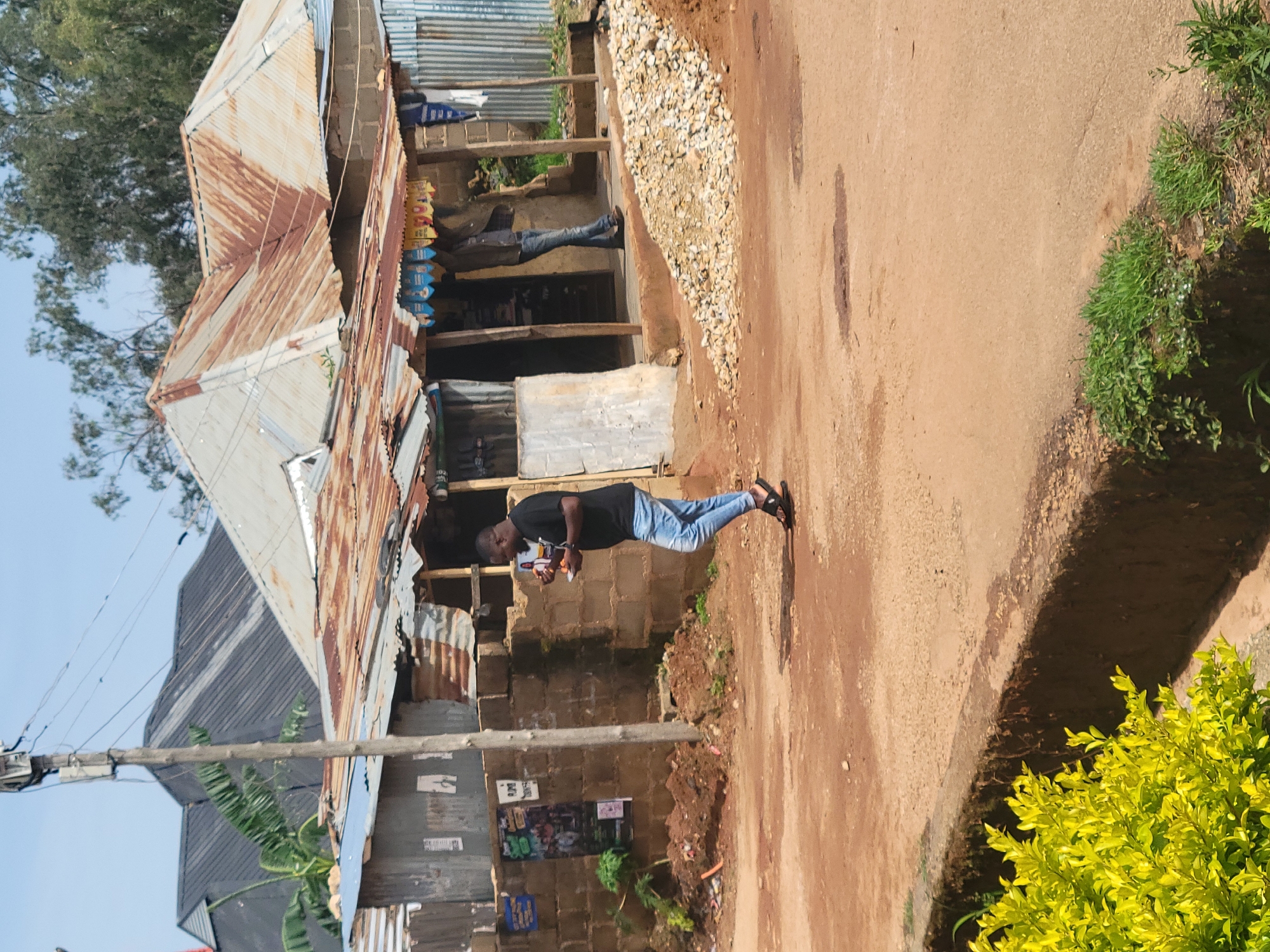 A man in a black t-shirt and light blue jeans walks on a dirt road in Jos, Nigeria, holding a small orange object in his right hand and a pamphlet in his left. He is walking past a dilapidated building constructed from cinder blocks and corrugated metal, with a rusted tin roof. The building appears to be a small shop or stall, with colorful decorations visible above the entrance. To the left of the building, a utility pole with wires stands prominently. The ground is a mix of reddish-brown dirt and scattered rocks, with a concrete curb and yellow-green foliage visible in the foreground. In the background, more buildings with dark, angled roofs and trees can be seen. The sky is clear and blue.