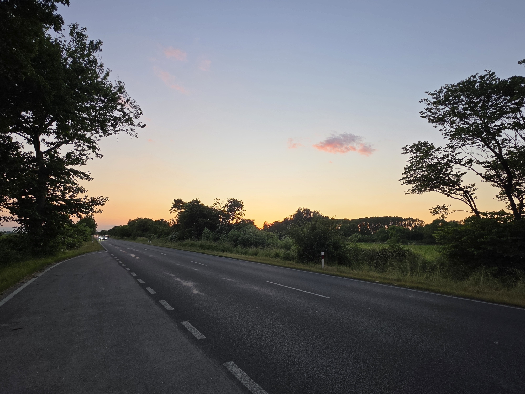 The image depicts a two-lane asphalt road cutting through a rural area. On either side of the road are trees and vegetation. The sky is illuminated by a sunset, with hues of orange and pink near the horizon transitioning to a light blue higher up. The road has white dashed lines separating the lanes, and a solid white line on the side closer to the viewer. A red and white pole marker is visible on the right side of the road. A car with headlights on can be seen further down the road to the left. Based on the user's specification, the location is Cheshnegirovo, Bulgaria.