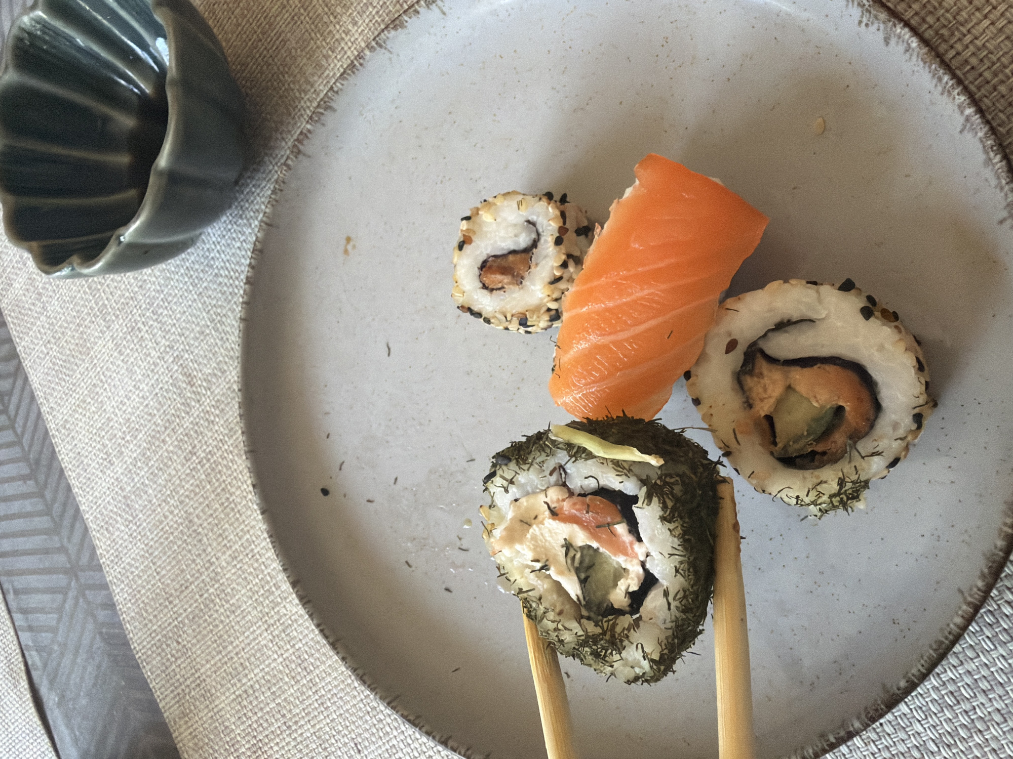 A close-up, overhead shot shows a plate of sushi. The plate, a speckled ceramic, is on a textured beige placemat, which is on a table with a patterned surface. In the upper right corner, a small, dark green, ribbed dish contains a dark liquid, likely soy sauce. On the plate, there are three pieces of sushi. Two are maki rolls, one with a dark green herb coating and what appears to be cream cheese and salmon inside, and another smaller roll coated in sesame seeds with a dark filling. Between the rolls is a slice of raw salmon. Two wooden chopsticks are holding one of the maki rolls. The scene is indoors, and the lighting suggests natural daylight.