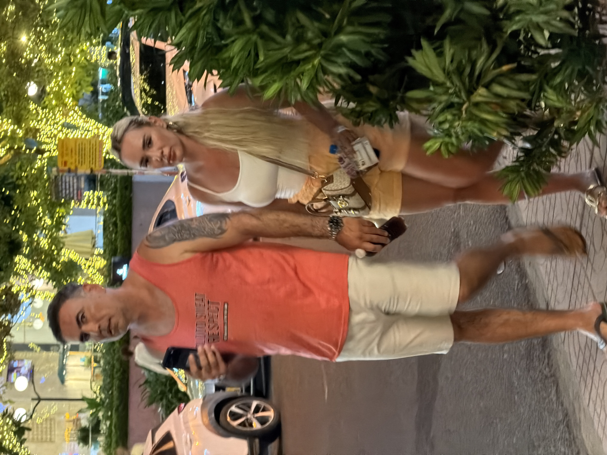 In Nha Trang Ward, Vietnam, at night, a man wearing a coral-colored tank top with "BLOOD SWEAT RESPECT" printed on it and white shorts walks alongside a woman with long blonde hair. She is wearing a white tank top and yellow shorts. The man holds a black smartphone in his left hand and a brown bottle in his right. The woman carries a small, patterned crossbody bag. They are walking on a sidewalk lined with trees illuminated by strings of yellow lights. Cars are visible in the background, along with buildings and other pedestrians.
