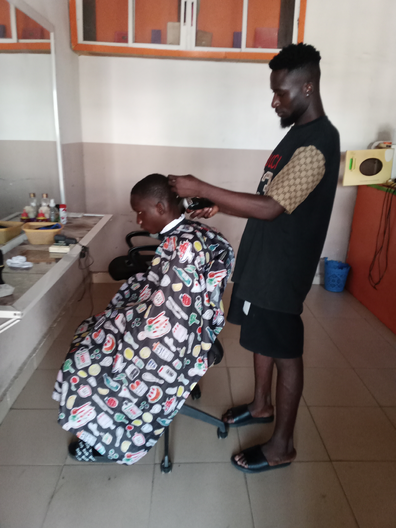 In a barbershop in Jalingo, Nigeria, a barber is trimming the hair of a young boy. The barber, a man with dark skin and a beard, wears a black t-shirt with beige checkered sleeves. He is using an electric clipper to cut the boy's hair. The boy, wearing a black cape with colorful cartoon food designs, sits in a barber chair. The shop has tiled floors and walls, with a mirror reflecting a counter filled with bottles and grooming supplies. The overall lighting suggests it is daytime.