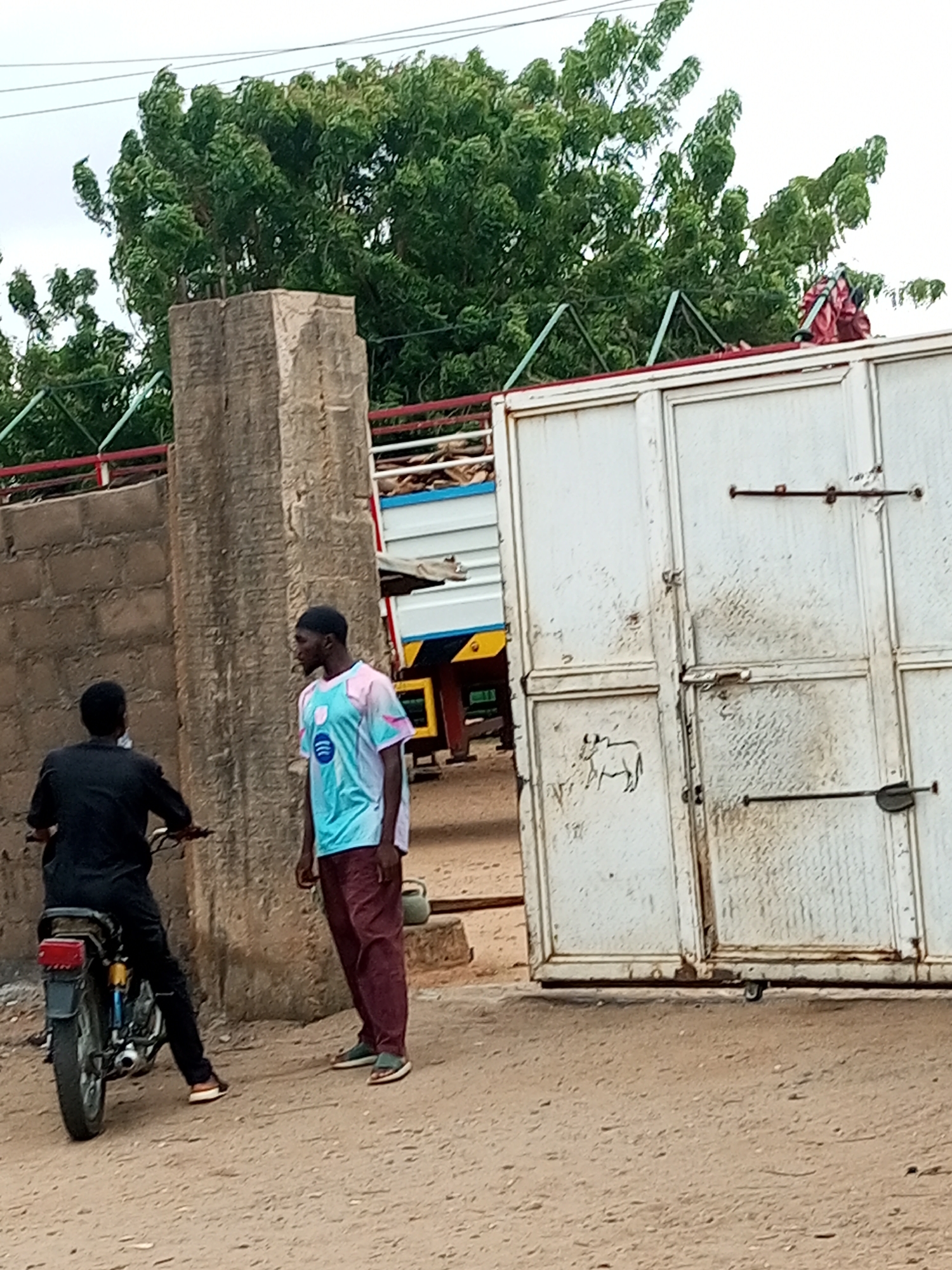 In the city of Tumfure, Nigeria, a dirt road leads to a large, weathered metal gate, possibly for a compound or industrial area. To the left of the gate, a textured, light-colored concrete pillar stands, with a brick wall extending from its base. In front of the pillar and wall, a man is seated on a motorcycle, facing away from the viewer and towards the gate. He is dressed in a black outfit. To the right of the pillar, another man stands, facing the motorcycle rider. He is wearing a turquoise and pink patterned shirt and maroon pants. Behind the men and the gate, a large truck with a white trailer is visible, with trees and foliage in the background under an overcast sky. The scene appears to be an outdoor, everyday setting in a Nigerian locale.
