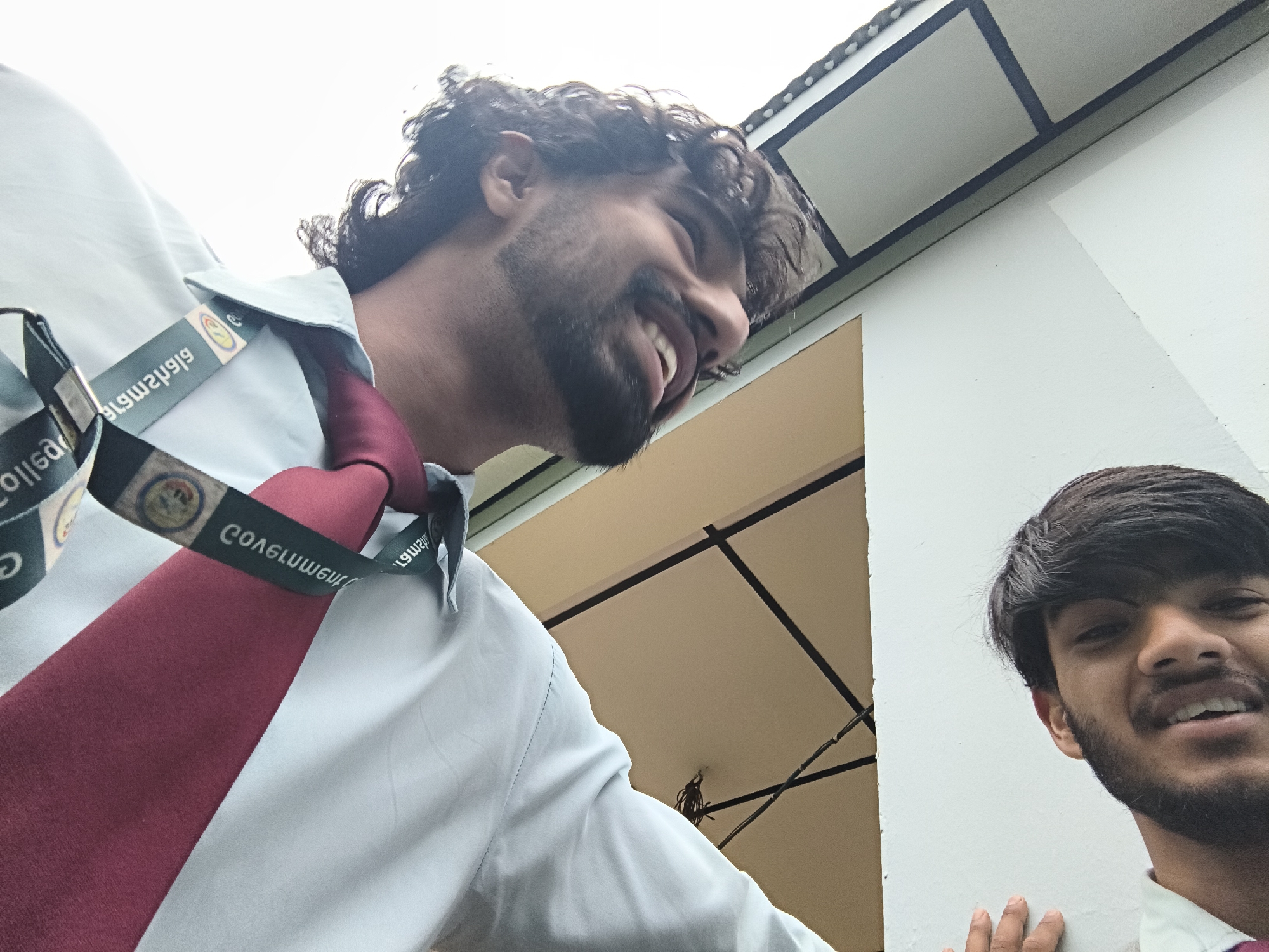 Two young men, dressed in white shirts and red ties, are captured in a low-angle selfie taken outdoors. The man on the left, with curly dark hair and a beard, is laughing. He is wearing a light blue shirt with a maroon tie and a lanyard with the inscription "COLLEGE" visible. The man on the right, with dark hair and a beard, is also smiling. They are positioned in front of a building with a light-colored exterior and an awning with dark trim. The overall setting appears to be a college campus or a similar institutional environment in Dharamshala, India, indicated by the presence of the building and the uniform attire of the individuals.