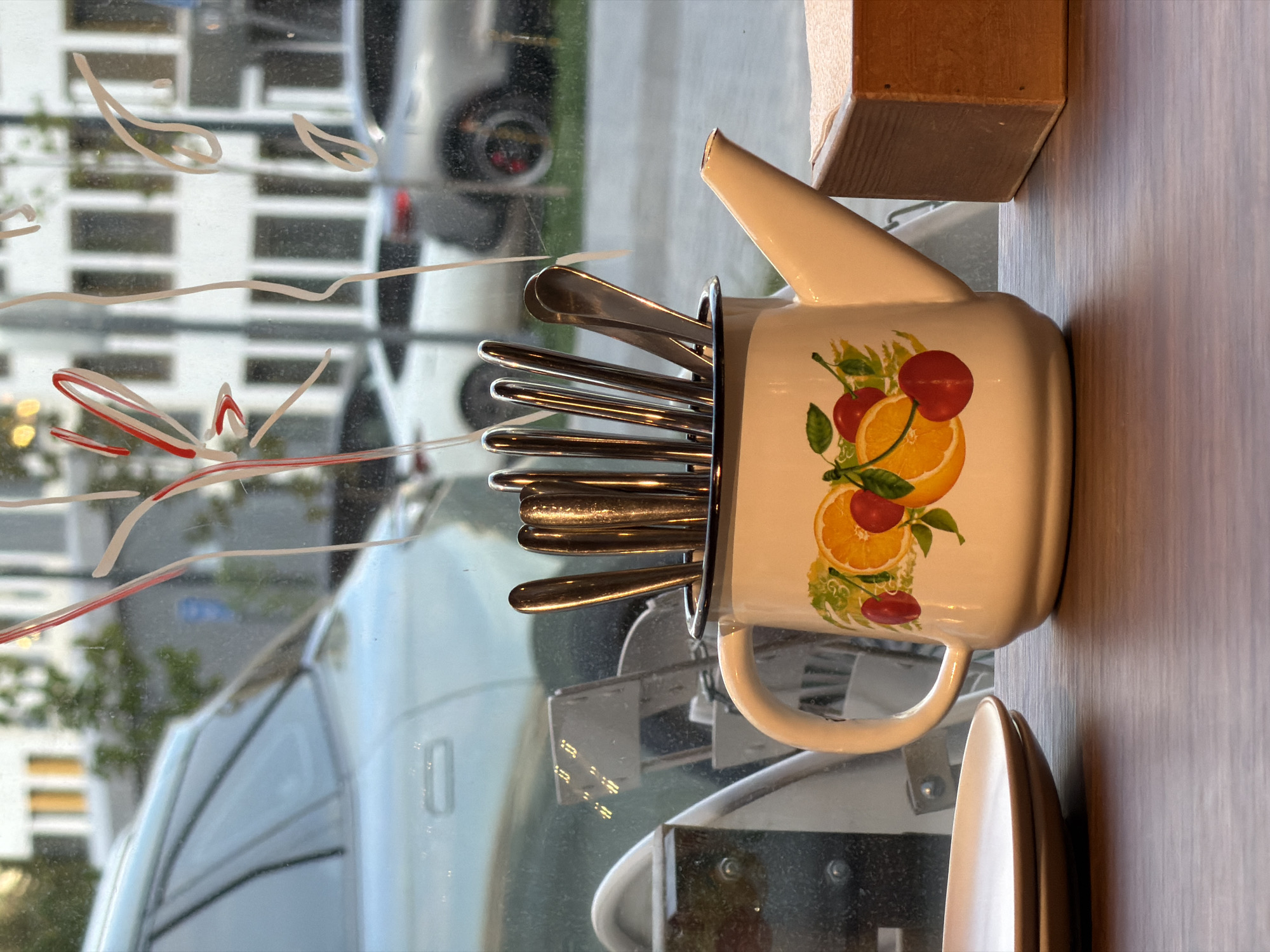 The image shows a still life arrangement on a counter. The main object is a white enamel pitcher with a spout, adorned with a pattern of oranges and cherries. It is filled with silverware, including spoons and forks. To the side of the pitcher is a wooden box. The background is a window with streaks of writing or markings on the glass, obscuring the view of the exterior. Through the window, one can discern parts of buildings and cars, indicating an urban setting. There is no visible indication of Izhevsk, Russia in the image.