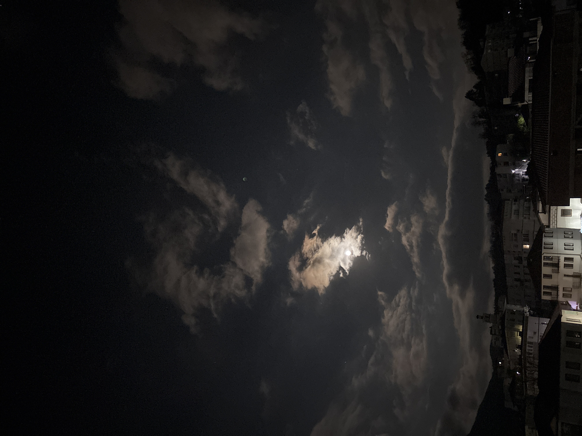 The image depicts a night scene in what appears to be a residential area. The sky dominates the view, featuring a mix of clouds partially obscuring the moon, which is the primary light source. The clouds vary in thickness and density, some allowing the moon's light to shine through more brightly than others.
On the right side of the image, apartment buildings are visible. The buildings are lit by streetlights and lights from inside the apartments, creating a contrast with the dark sky. The architecture suggests a city setting. Although I don't have enough information to confirm that it is in the city of Zlatograd, Bulgaria.