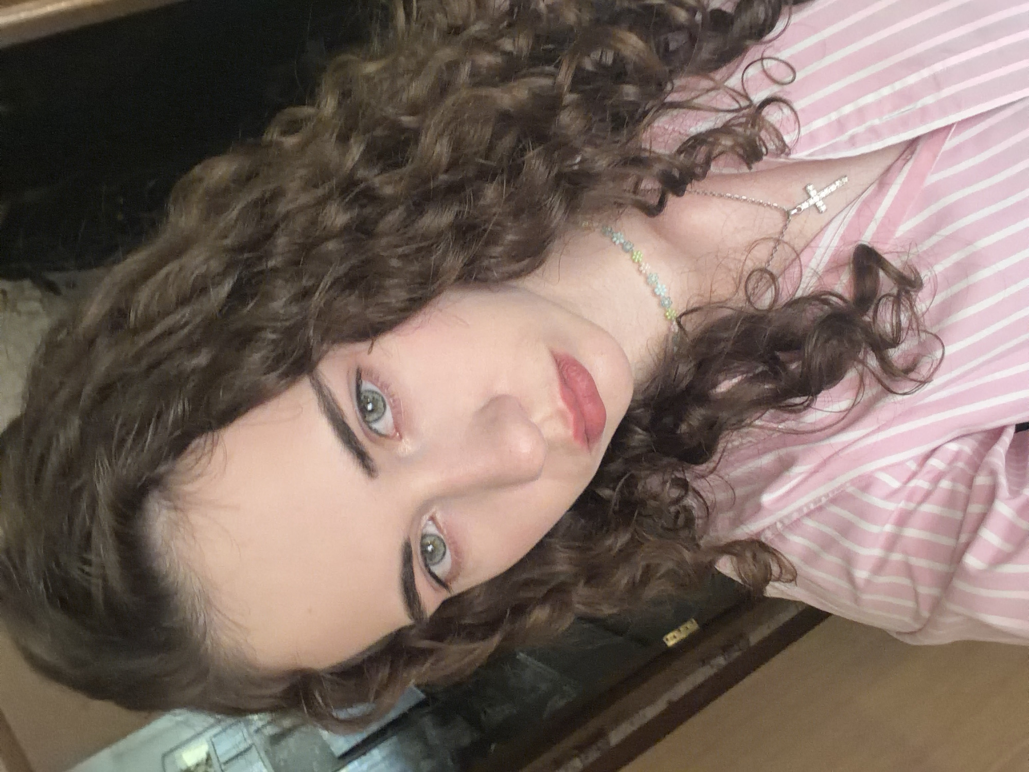 A young woman with curly brown hair and green eyes is looking into the camera. She is wearing a pink and white striped shirt and a necklace with a cross pendant. The background is blurred and appears to be an indoor setting. There is no indication of the specific location within the city of Zavodoukovsk, Russia.