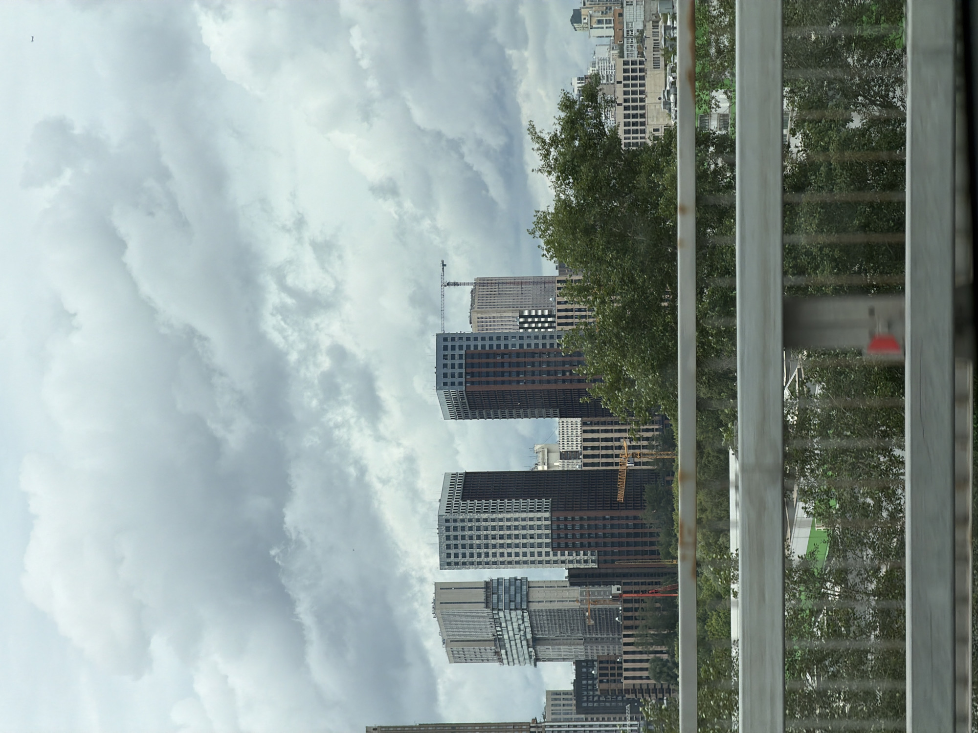 This is a medium-shot, eye-level, outdoor photograph of a cityscape under a cloudy sky, taken from a bridge with a metal railing in the foreground. The railing is silver-colored and appears weathered, with horizontal bars and vertical supports. Beyond the railing, a dense collection of modern high-rise buildings dominates the midground. The buildings exhibit a variety of architectural styles and materials, including glass, concrete, and brick facades in shades of grey, white, and brown. Some of the buildings appear to be under construction, indicated by the presence of yellow construction cranes. Green trees and foliage are visible between and in front of the buildings, suggesting a park or green space within the urban environment. The sky is filled with heavy, grey clouds, obscuring any direct sunlight and giving the scene a somewhat overcast and muted atmosphere. The overall impression is one of a contemporary urban development, likely in a district of Moscow undergoing modernization or expansion. The perspective suggests the photo was taken from an elevated position, such as a road or pedestrian bridge.