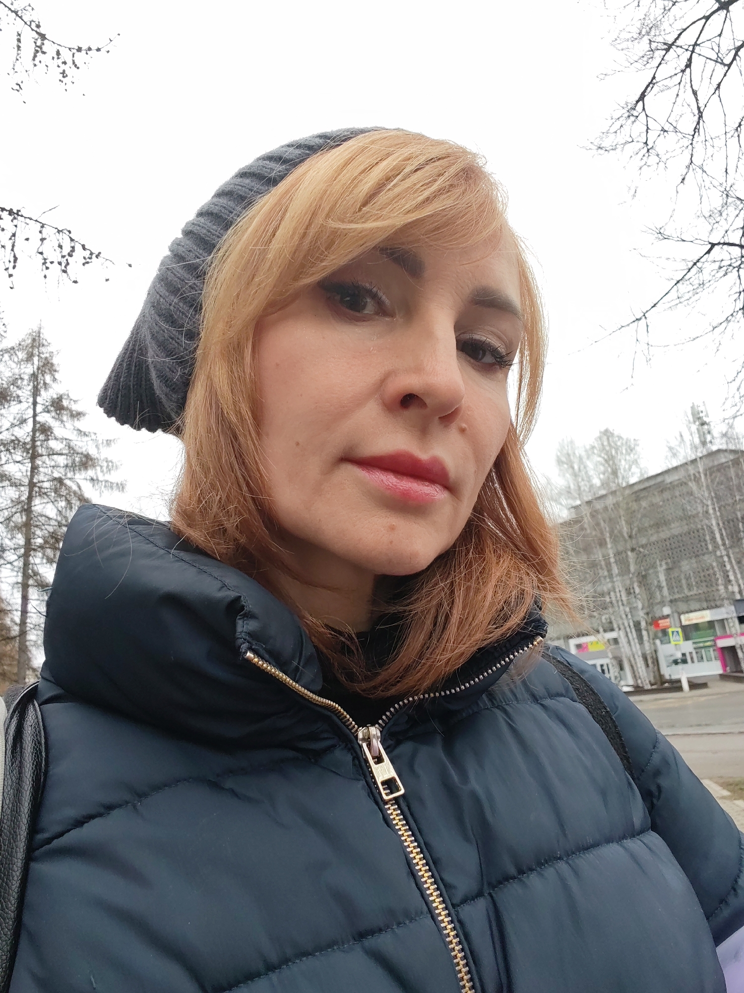 A woman with red hair, wearing a gray knitted hat and a dark blue puffer jacket with a gold zipper, is looking at the camera. The background features a city street with buildings, trees, and a cloudy sky. The image was likely taken in the city of Chaikovsky, Russia.
