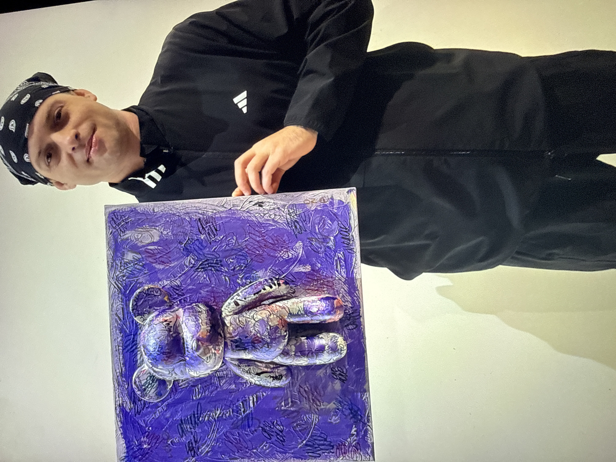 A man wearing a black bandana, a black Adidas jacket, and black pants holds up a piece of art. The artwork is a purple and silver bear figurine attached to a purple, textured background. The background has scribbled text in black and white. The man is smiling and looking at the camera. The background is a plain, light-colored wall with a shadow cast on it. The image appears to have been taken indoors, possibly at an art exhibition. The location is not specified in the image or the provided crops.