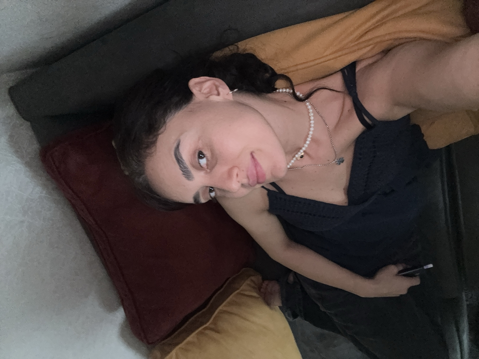 A young woman is lying down on a couch. She is wearing a dark blue top and dark pants, and a pearl necklace. She is holding a phone in her left hand. The couch is dark gray and there are several pillows behind her, one red and one yellow. The lighting is dim. The image is a selfie taken from a slightly overhead angle. The woman appears to be in a domestic setting, possibly a living room or bedroom. The specific location in Tbilisi, Georgia cannot be determined from the image alone.