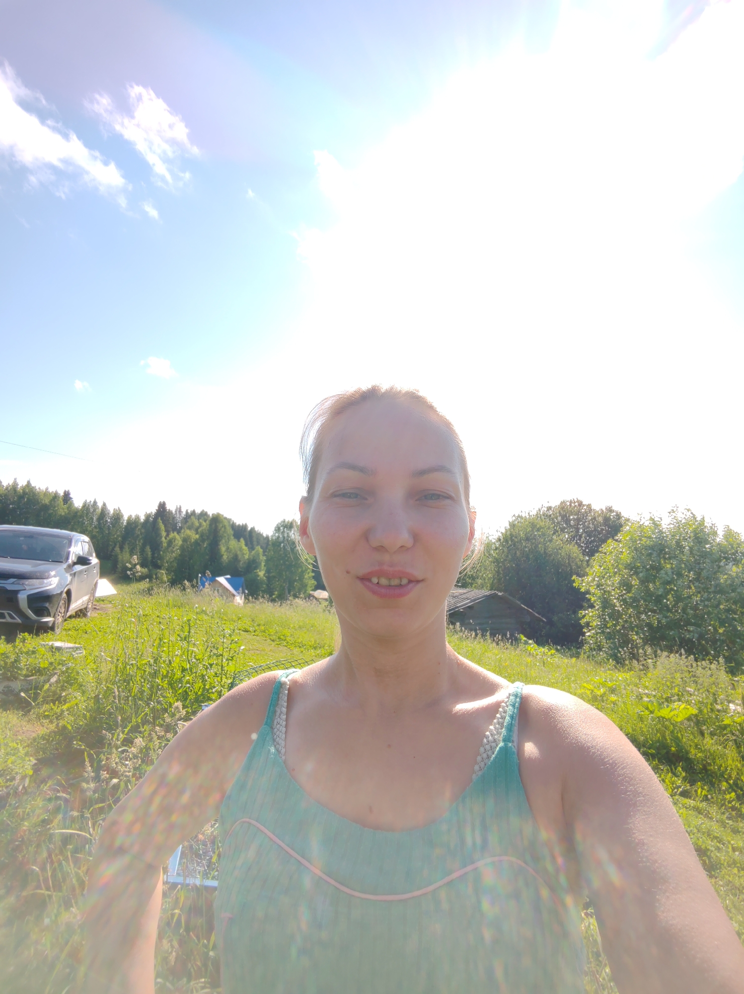 This is a medium shot, selfie-style photo taken outdoors on a sunny day. The subject, a woman with blonde hair and fair skin, is positioned in the foreground, her face close to the camera. She is wearing a light blue tank top with pink trim and braided straps. Her expression is neutral, with a slight smile.
In the background, a dark-colored SUV is parked on the left side of the image. Further back, a wooded area with green trees stretches across the horizon. There are also glimpses of what appear to be small buildings or structures scattered among the trees and in a grassy field in the middle ground. The sky is bright blue with scattered white clouds and a strong sun flare at the top right of the image, indicating the sun is high and bright. The environment suggests a rural or semi-rural setting, possibly a dacha or country property, consistent with locations like Чухлэм, Russia. The overall lighting is bright and airy, with some lens flare creating colorful streaks across the image, particularly on the subject's arms and upper body.