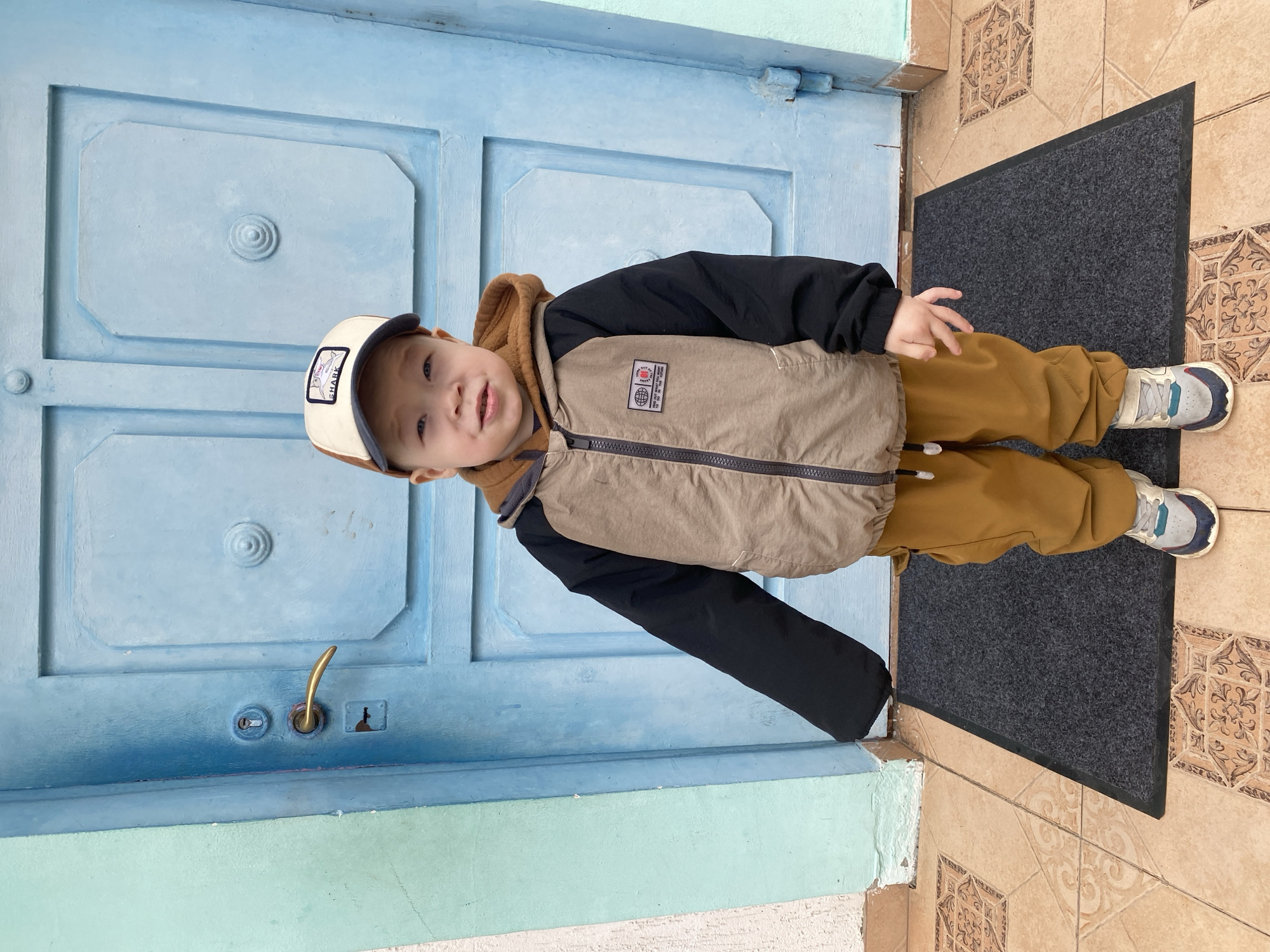 A young child stands in front of a light blue door with a black doormat on a tile floor, possibly in Sochi, Russia. The child wears a two-toned jacket (black and beige), brown pants, and sneakers, with a white and tan cap. The door is paneled, and the tile floor is decorated with patterned squares.
