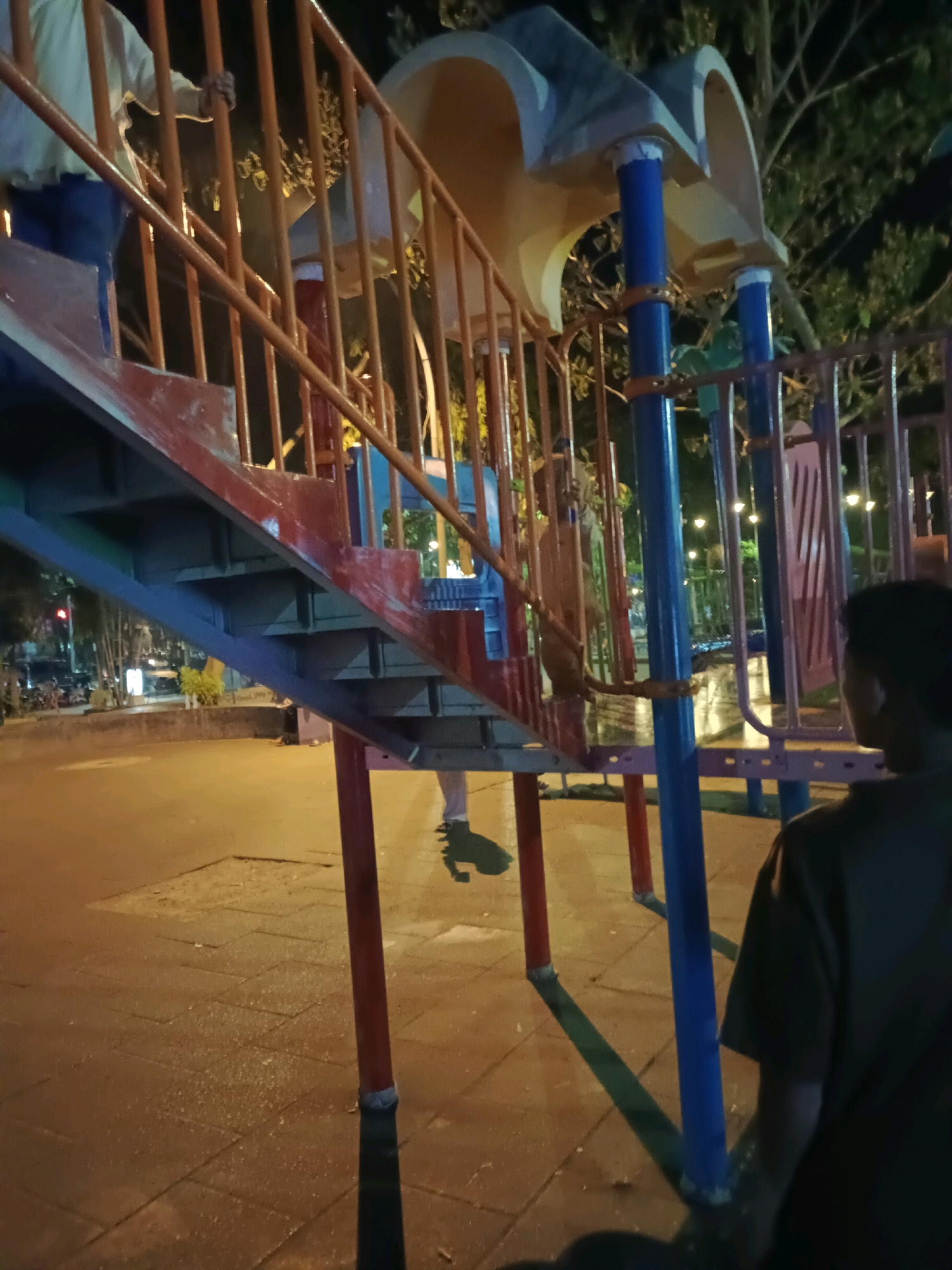 The image depicts a playground structure at night in Lamongan, Indonesia. The playground features a metal staircase with orange railings and red accents, leading up to a yellow and gray plastic slide. The ground surface appears to be paved with interlocking bricks and is wet, suggesting recent rain.
In the foreground, a person wearing a dark t-shirt is visible from the back, looking towards the playground. Further into the scene, on the left side, part of another person is seen on the stairs, wearing a light-colored shirt and blue pants. Another person, partially obscured by the playground structure and the fence, appears to be standing behind the railings. The background shows trees and streetlights, indicating an urban environment. Vehicles and a traffic light are also visible in the distance, suggesting the playground is located near a road. The overall lighting is dim, with the playground structure being illuminated by artificial lights.