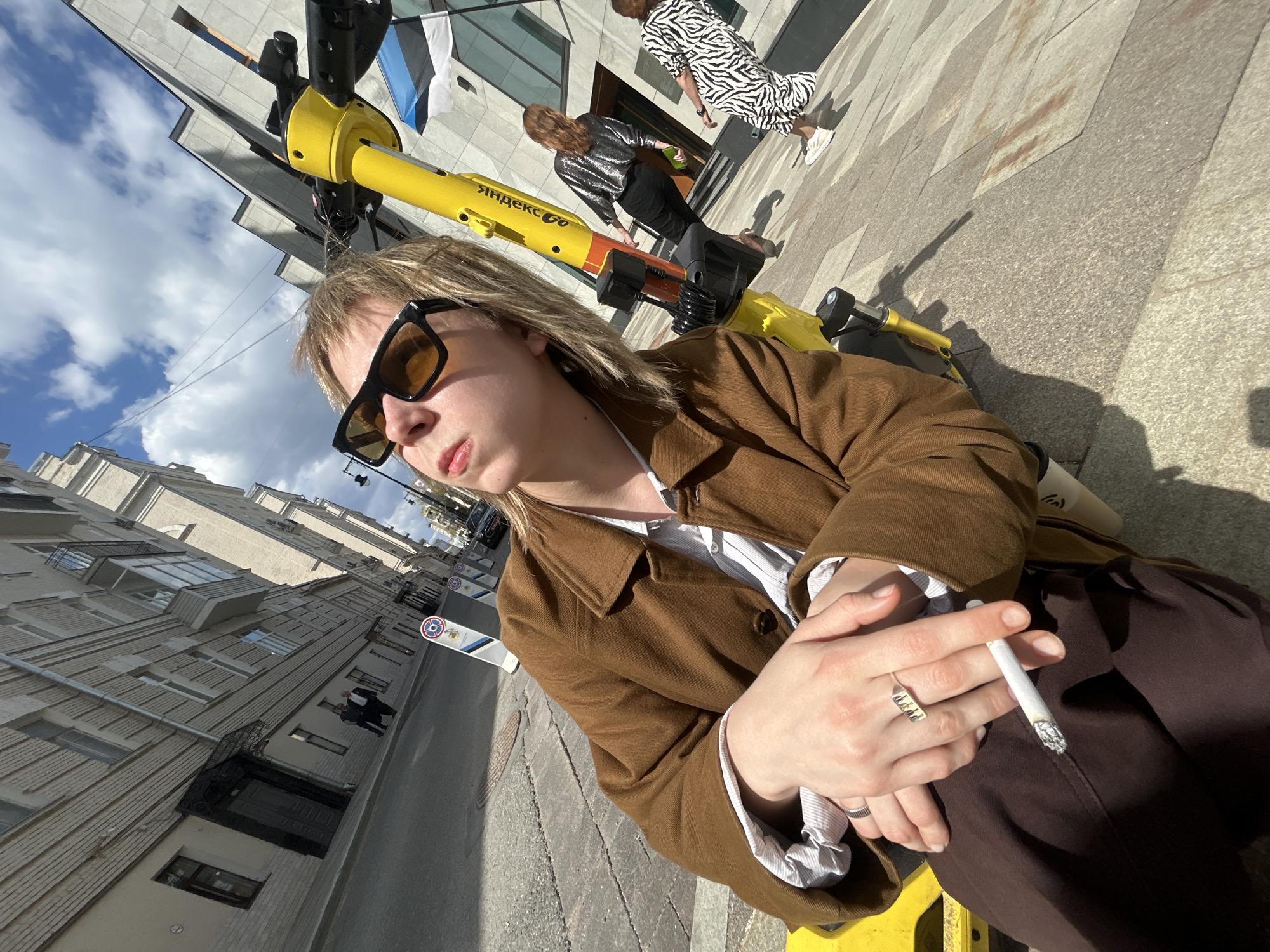 A woman wearing sunglasses and a brown jacket is sitting on a yellow Yandex Go electric scooter in an urban environment. She is holding a lit cigarette in her right hand. Behind her, two other people are walking. The woman in front is wearing a sequined top and black pants, while the person behind her is wearing a zebra-print dress. The background includes buildings, a street, and the Estonian flag. The image appears to be taken at an angle from the ground, creating a slightly tilted perspective.