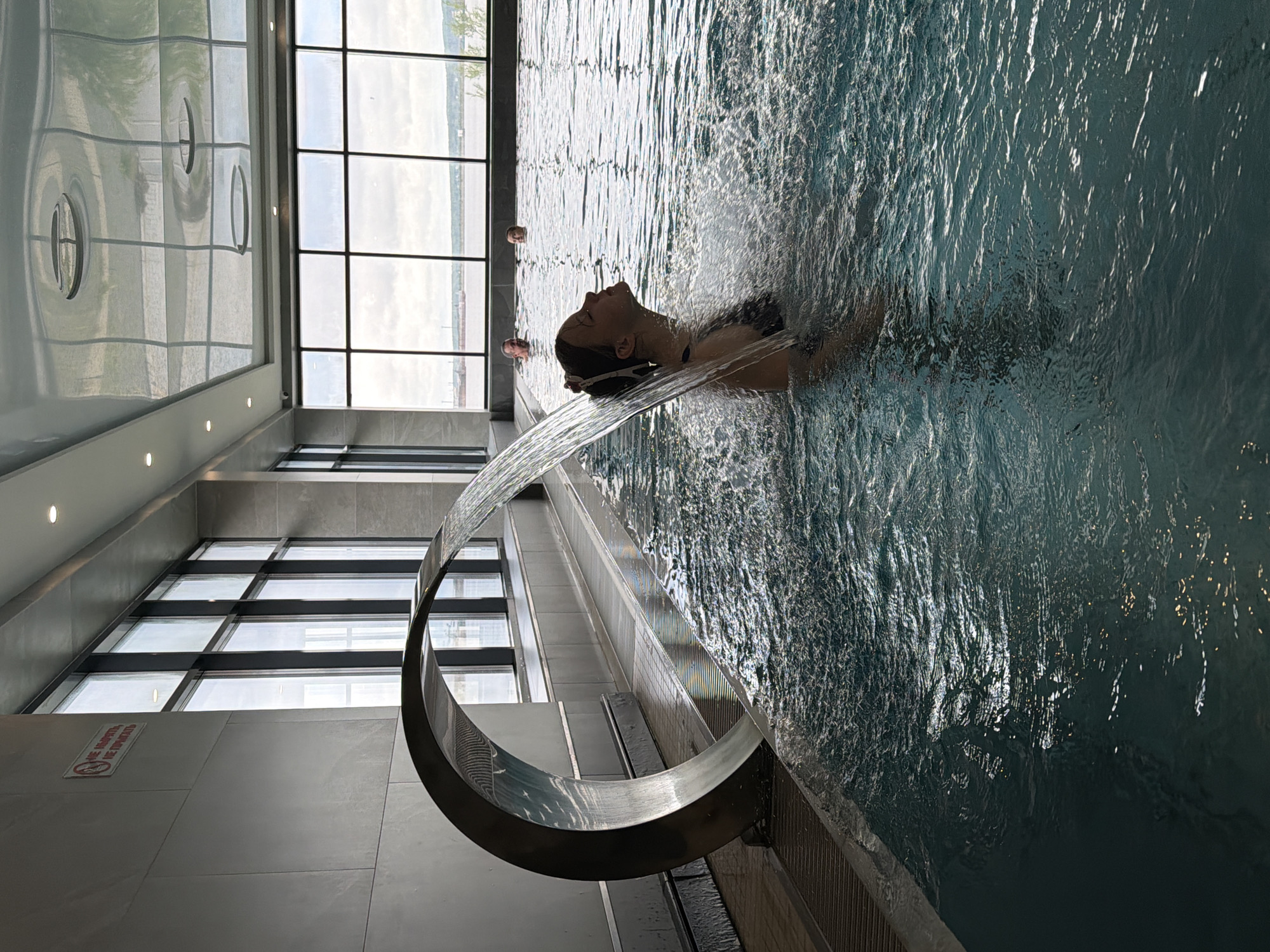 A woman is in a swimming pool with a water feature in the city of Chaikovsky, Russia. She is positioned under a stream of water flowing from a crescent-shaped metal structure, with her head tilted back and eyes closed. The pool is indoors, with large windows overlooking an outside area. Two other people are visible at the far end of the pool. The architecture is modern, featuring sleek lines and a combination of tiled and reflective surfaces. A sign is visible on the wall.