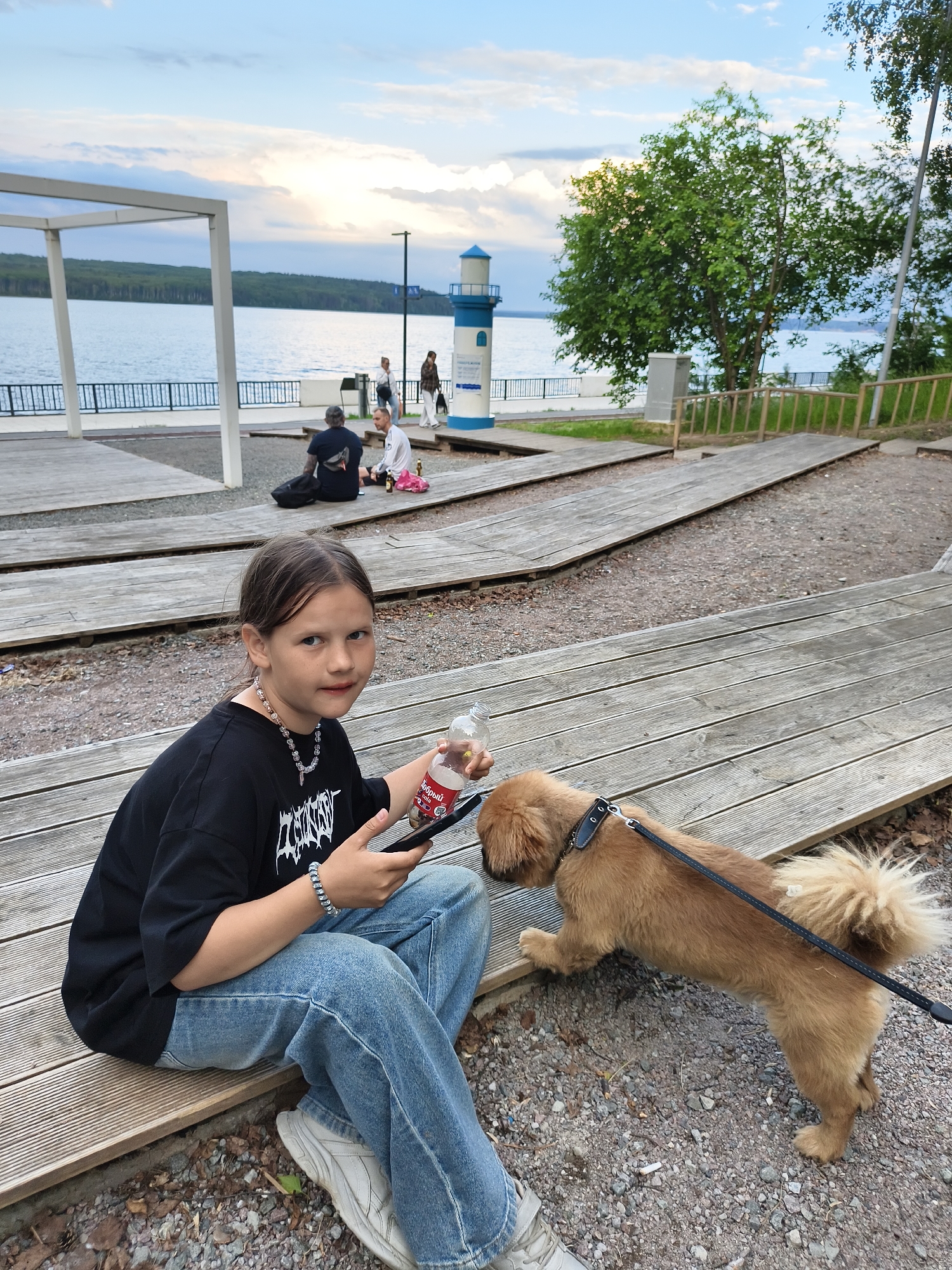 A young girl with dark hair and blue eyes, wearing a black t-shirt with a white graphic and blue jeans, sits on a wooden bench. She holds a clear plastic bottle with a red label and a black smartphone in her hands. A small, fluffy brown dog on a black leash stands next to her, sniffing the ground. The girl looks towards the camera with a neutral expression.
The setting appears to be an outdoor recreational area with a body of water in the background, likely a lake or river. Several other people are visible in the distance, some sitting on benches and others walking. A small lighthouse-like structure stands near the water. The sky is overcast with patches of blue, suggesting either early morning or late afternoon. The ground is covered in gravel and fallen leaves, and wooden walkways or seating areas are scattered throughout. The overall atmosphere seems relaxed and casual.
The red label on the bottle has text, but it is not clearly legible. There is also text on the girl's t-shirt, appearing to be a stylized white graphic. The time of day appears to be late afternoon or early evening, given the lighting. The location context indicates Chaikovsky, Russia.