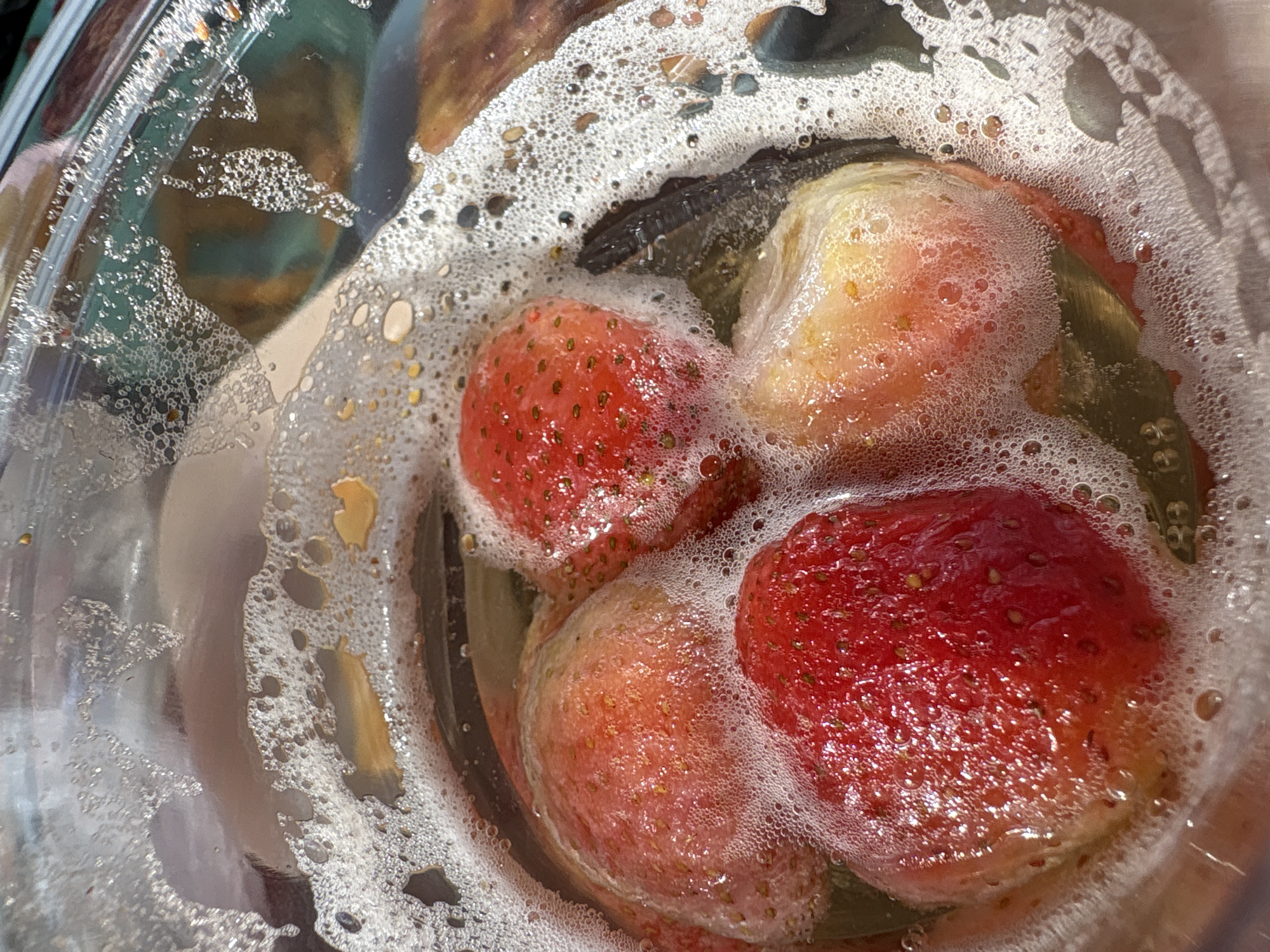 The image shows four strawberries soaking in a clear glass bowl filled with soapy water. The strawberries are mostly red, with some variations in color. The soapy water creates a ring of bubbles around the strawberries. There are no people in the scene. The image does not suggest the location is in the city of Chaykovsky, Russia.
