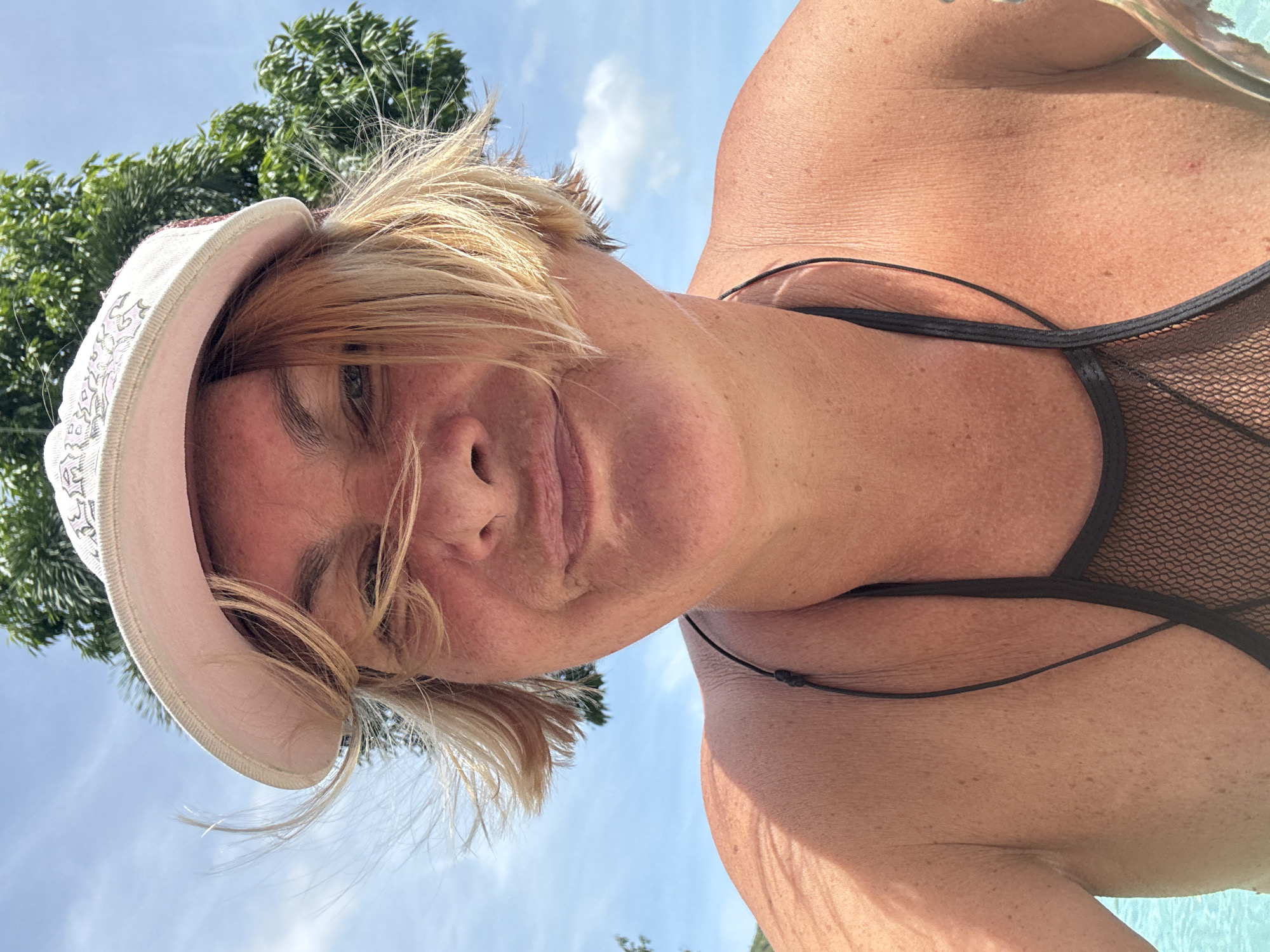 A low-angle, close-up photograph shows a woman wearing a white cap and a black swimsuit. The setting appears to be outdoors, likely in a natural or resort environment. The woman has fair skin and blonde hair. In the background, there is a tree, a blue sky, and white clouds. The location is บ้านบางรักษ์, Thailand.