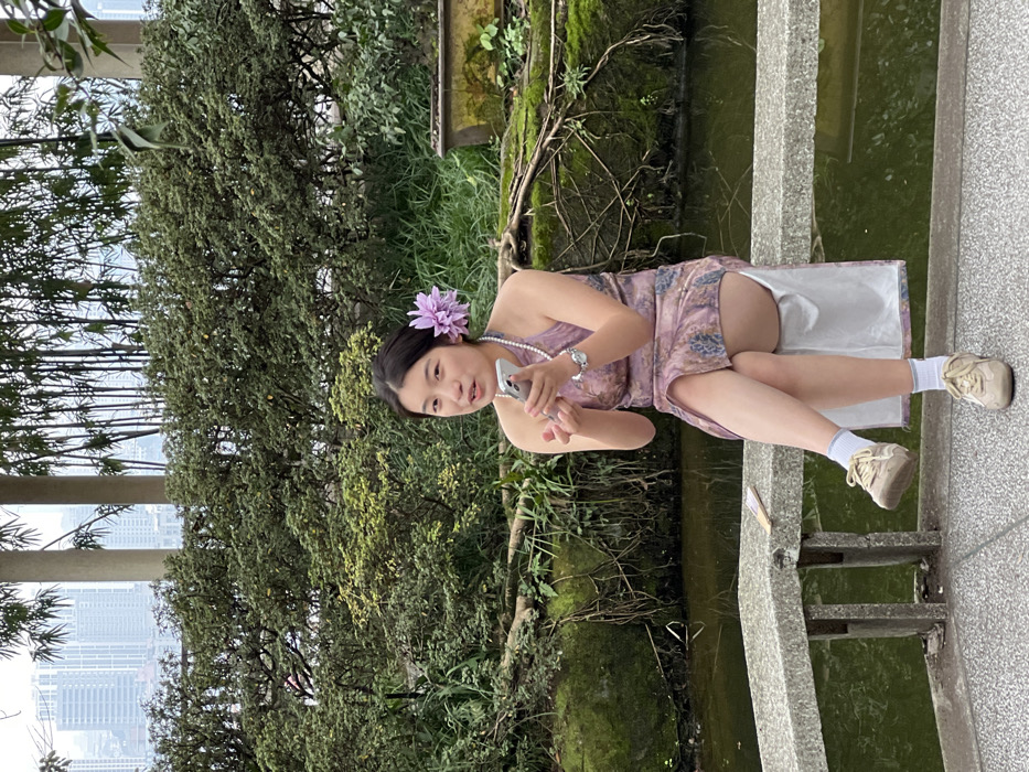 A young woman in a floral-patterned dress and white socks sits beside a canal. She has a purple flower in her hair and is holding a cell phone. She is wearing a pearl necklace and a watch. A lush green forest and a distant cityscape are visible. The canal walls are stone and covered in moss.