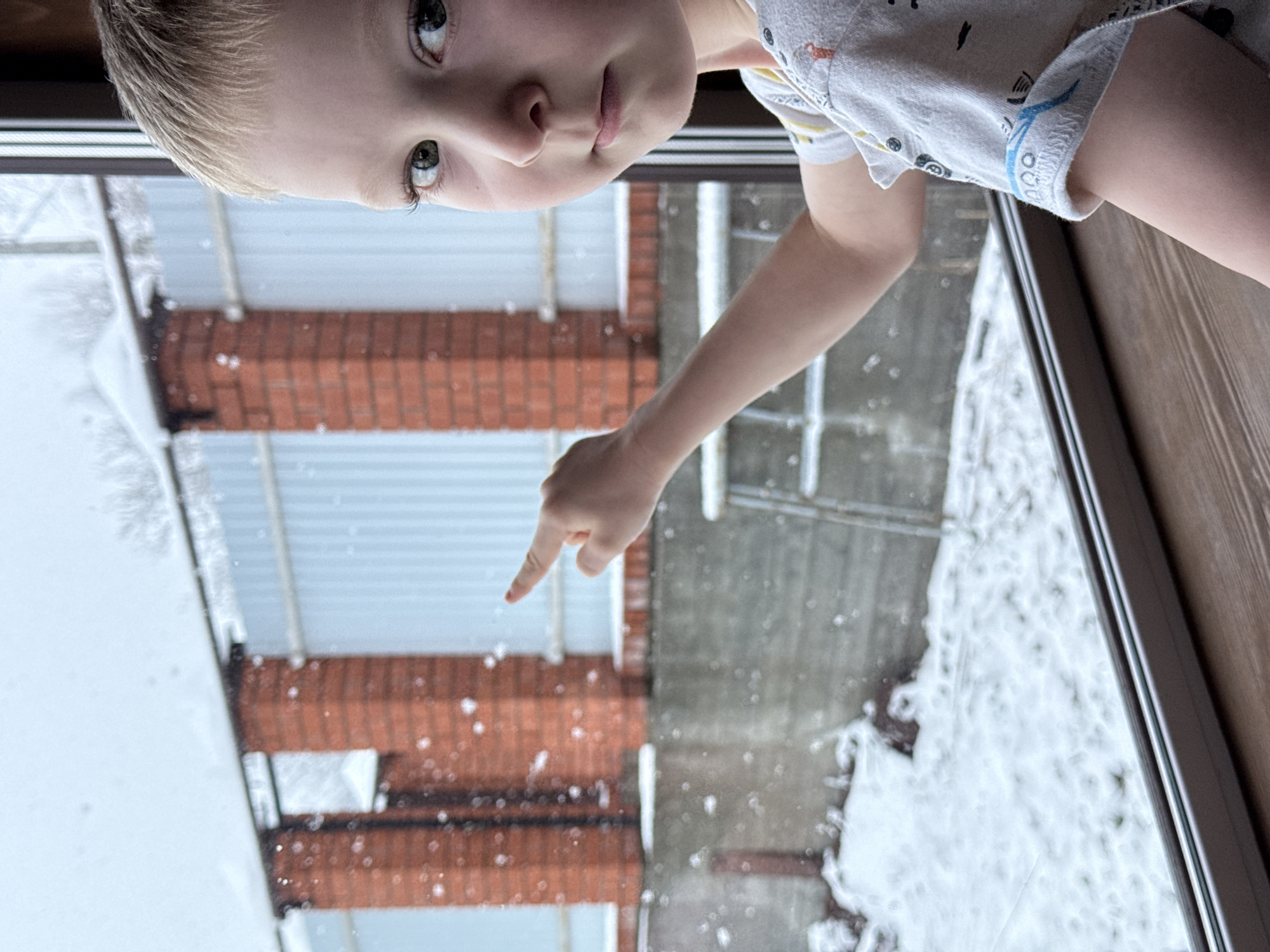 A young child is seen from a high angle, possibly looking out a window. The child has blonde hair and is wearing a light-colored shirt with a pattern. Their arm is extended with a finger pointing downwards. Outside the window, there is a scene of snow falling. A brick building with a corrugated metal roof is partially visible through the window. The falling snow is visible against both the building and the sky. The location is suggested to be Perm, Russia.