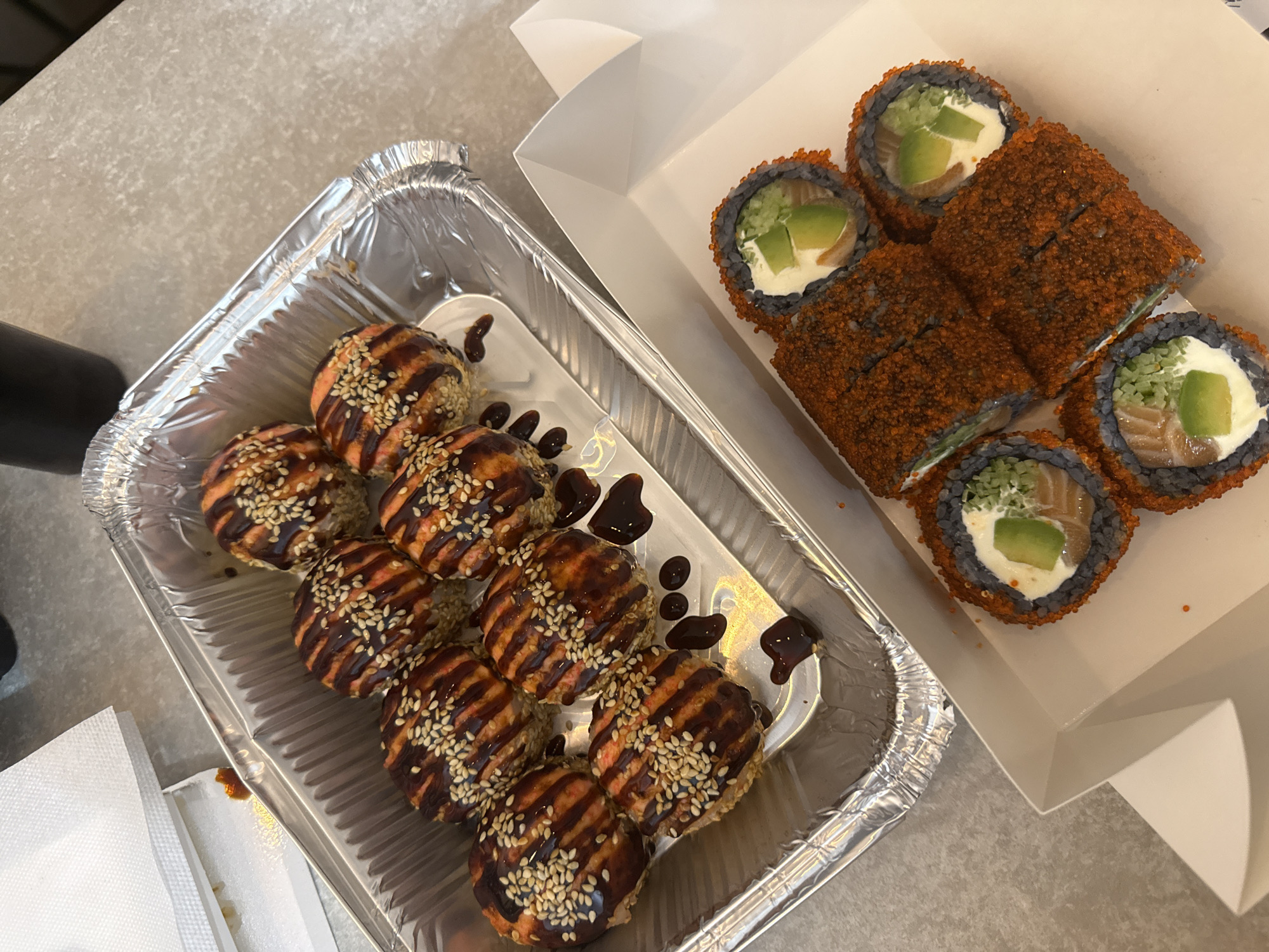 The image depicts two containers of sushi on a gray surface. The container on the left is a silver aluminum tray holding a dozen sushi rolls arranged in a triangular formation. These rolls are topped with a striped brown sauce and sesame seeds, with droplets of the same sauce decorating the tray around the rolls. To the right is a white cardboard container holding sushi rolls coated with orange roe. These rolls feature a dark gray nori wrap and are filled with cream cheese, salmon, and avocado. The setting appears to be indoors, possibly a restaurant or a home. The location is not evident from the image alone.