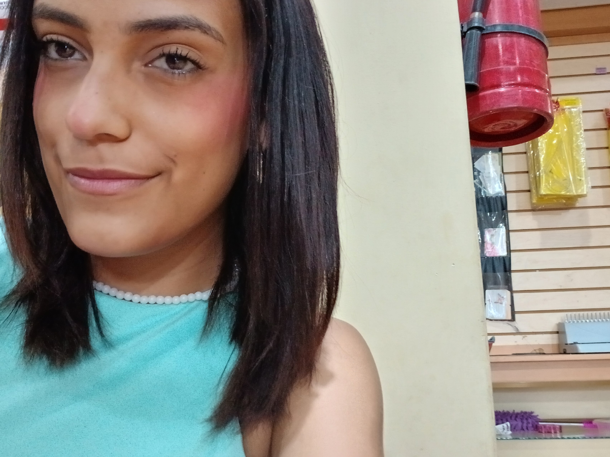 The media file is a close-up, self-portrait style photograph of a young woman with dark hair, looking directly at the camera with a slight smile. She is wearing a light blue top and a delicate white beaded necklace. Her makeup includes visible blush on her cheeks.
The background is out of focus but suggests an indoor setting with a beige wall on the left and a wall covered in horizontal wooden planks on the right. On the wooden wall, there are several items displayed. A large red object, possibly a fire extinguisher, is prominently visible hanging from the top. Below it are various items in yellow packaging and a display rack holding small cards or photographs. Further down, shelves hold a blue object with many thin vertical lines, and a purple duster-like object. The lighting appears to be artificial, with no indication of time of day or weather.
The overall mood is casual and direct, typical of a selfie. There is no visible text in the image. The location context provided indicates Juan Griego, Venezuela.