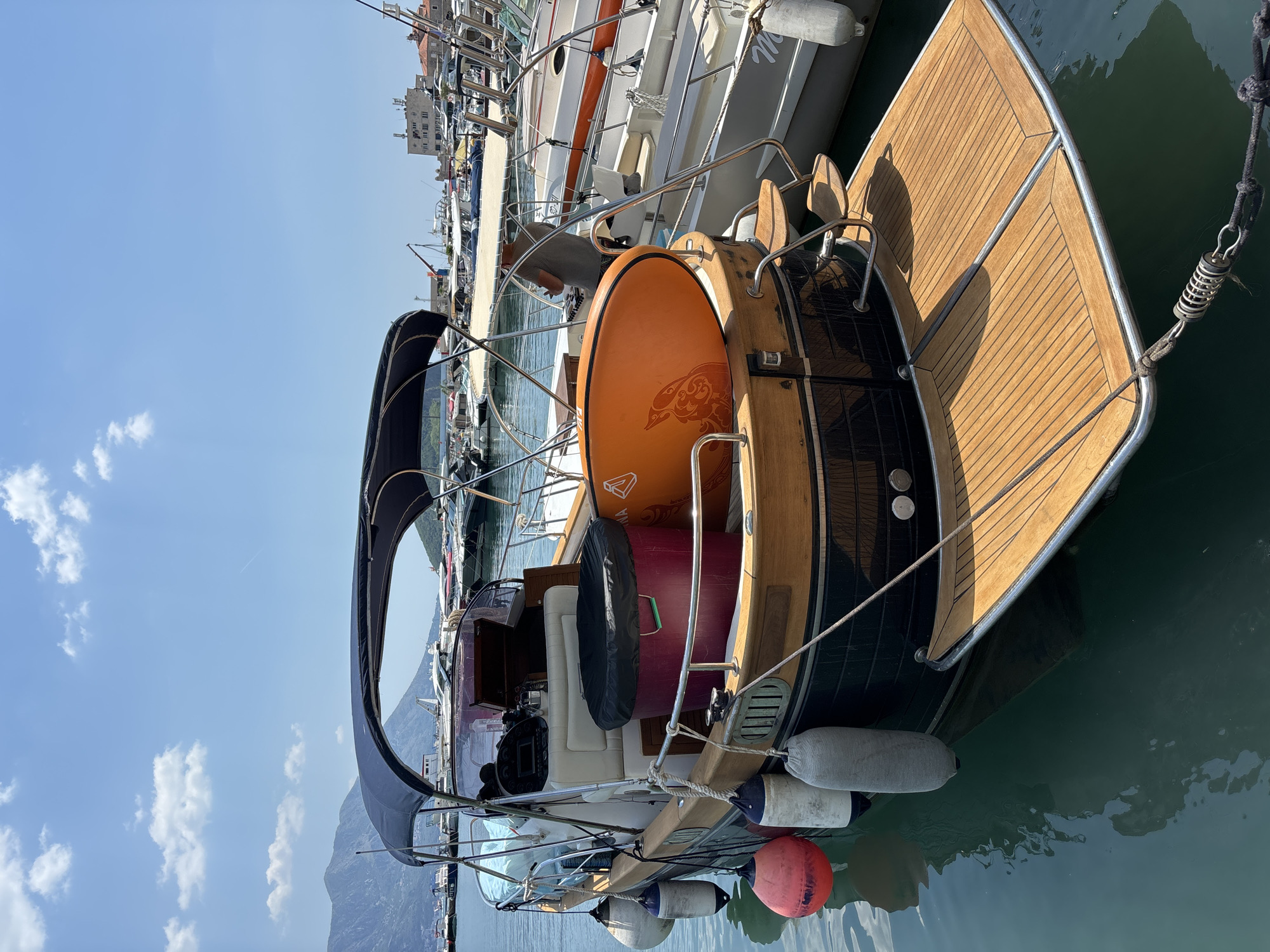 A motor yacht is moored in a marina in Budva, Montenegro, with a clear blue sky overhead and scattered clouds. The yacht has a dark hull and a wooden deck at the stern. It is equipped with a navy blue canopy over the cockpit. On the deck, an orange stand-up paddleboard with an orange design is leaning against a large, dark-lidded red container. Several buoys, two white and one red, are attached to the side of the yacht. In the background, other boats are docked, and a city skyline with buildings and mountains is visible. The water is calm and reflects the sky.