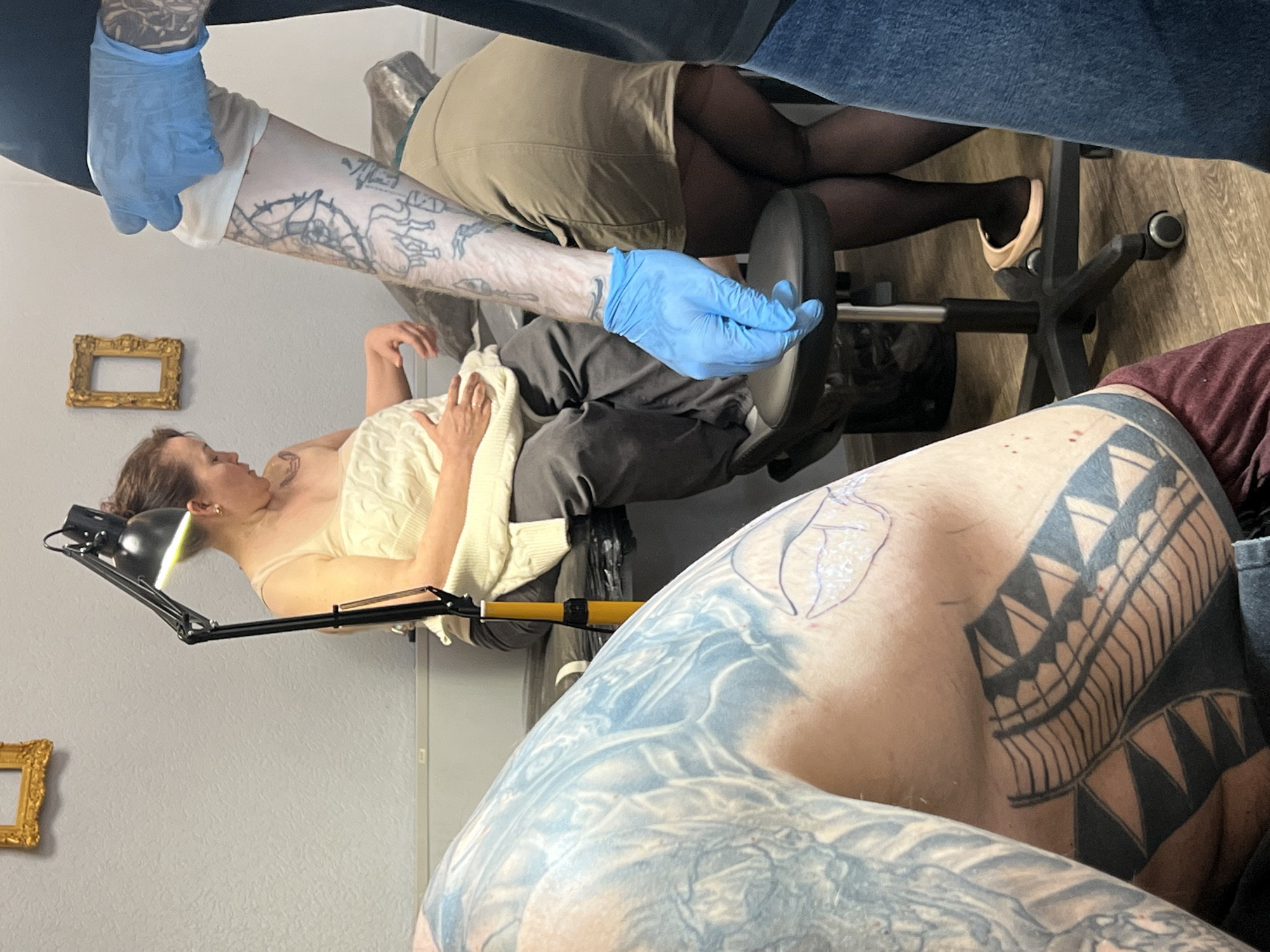 The image depicts an active tattoo session taking place in what appears to be a professional tattoo studio, with the photo rotated 90 degrees counter-clockwise.
A female client is lying on her back on a padded tattoo chair, her head resting on a cushion, with a black articulated desk lamp illuminating her. She wears a light-colored, possibly cream or white, knit top. Her left forearm, extended upwards, is being tattooed. This arm already features several outline tattoos, and a new, intricate blackwork design, resembling thorns or intertwined branches, is in progress.
A tattoo artist's arms are visible, heavily tattooed themselves, and wearing light blue nitrile gloves while working on the client's forearm. Another person, possibly an assistant or another artist, is partially visible in the background, seated on a wheeled office chair, wearing khaki shorts or a skirt and black tights. In the foreground, the heavily tattooed shoulder and upper arm of a third individual are prominent. This person's skin is adorned with extensive black tribal-style patterns and lighter blue/grey designs, with a fresh stencil for a new, organic or floral tattoo visible on their upper shoulder. They are wearing a dark red or maroon shirt.
The setting includes light grey walls, one of which has a golden empty picture frame hanging on it. The floor is made of wooden planks. The overall atmosphere is focused and professional, highlighting the detailed artistry of tattooing. The scene is consistent with a modern tattoo parlor, potentially located in Saint Petersburg, Russia. No discernible text is visible in the image.