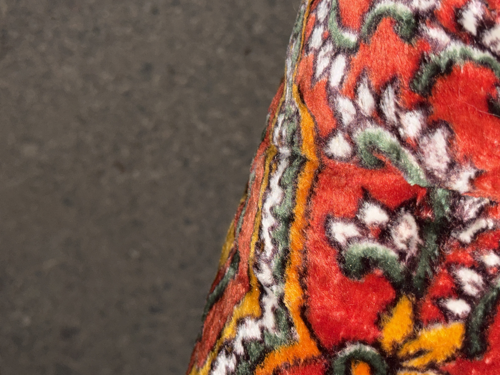 A close-up, macro shot shows a richly colored, plush fabric, possibly a rug or tapestry, with a vibrant red base. Intricate patterns of yellow, green, and white are woven into the fabric, creating a floral or paisley-like design. The texture appears soft and velvety. The fabric is angled upwards, with the upper portion of the image showing a blurred, dark grey, textured surface, likely asphalt or concrete, suggesting an outdoor urban environment. The context of Vostok, Kyrgyzstan is not visually evident from the provided image.